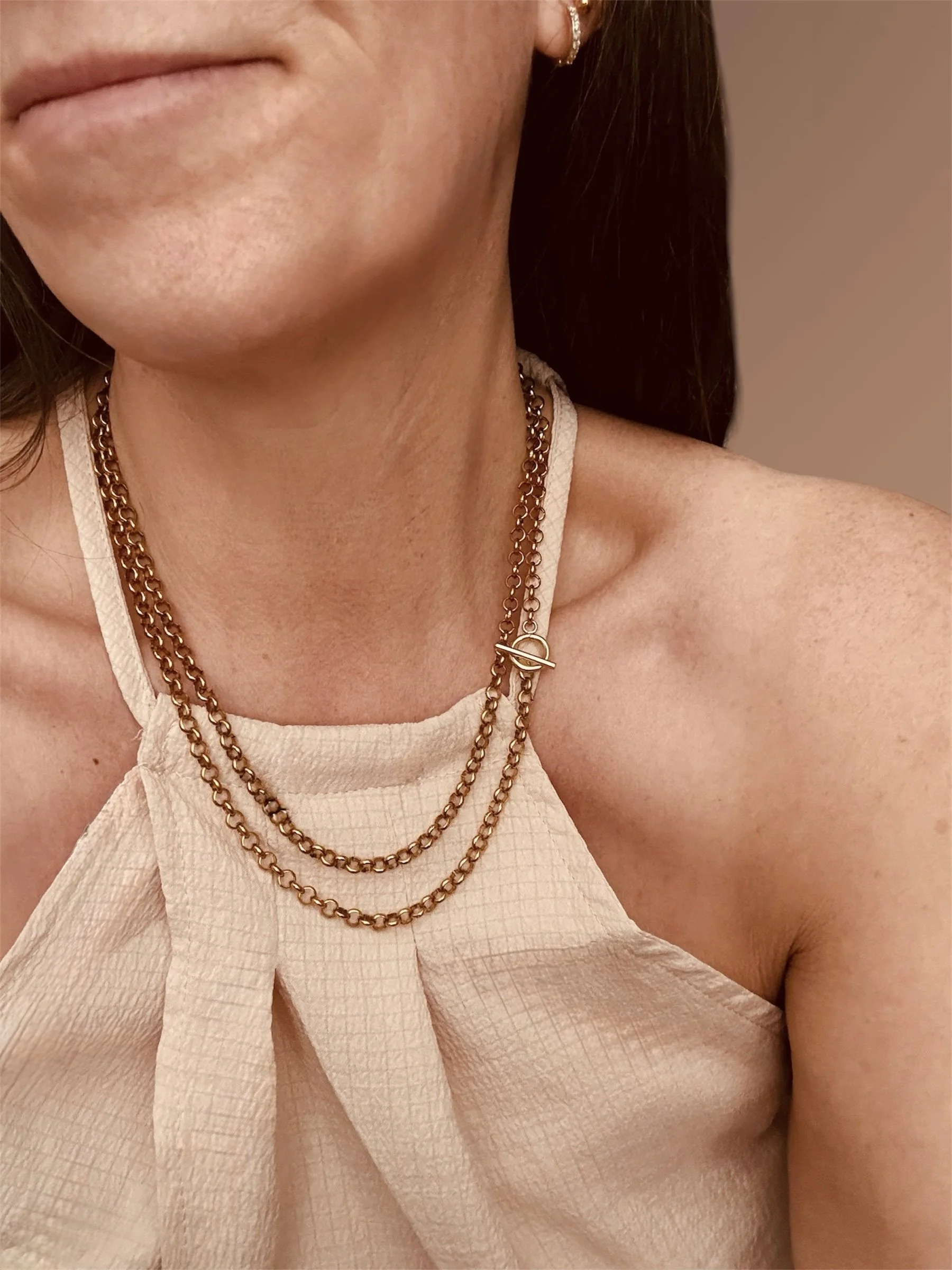 Close-up of a woman's lower face, neck, and shoulder, showing gold chain necklaces, a white textured top, and earrings, with a blurred neutral background.