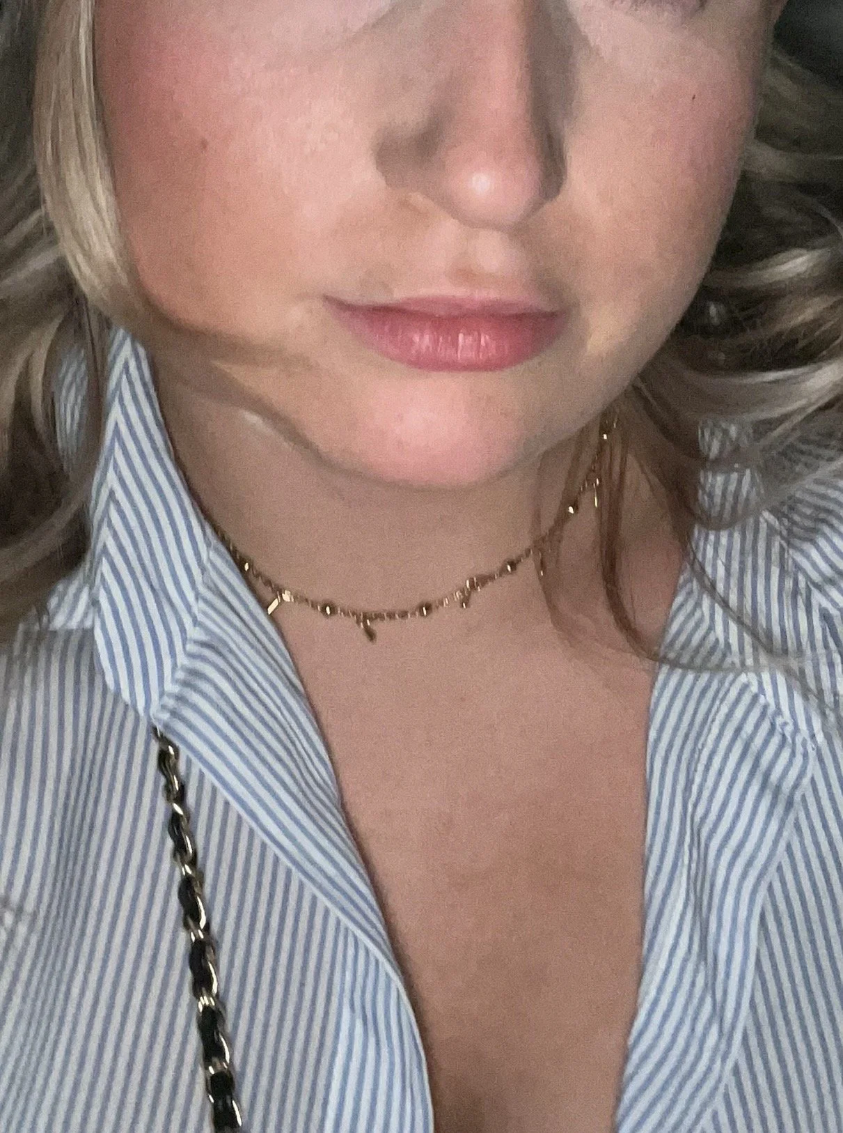 Close-up of a woman wearing a blue and white striped shirt, gold necklace with small charms, and a black and gold chain strap, with blonde and brown curly hair.