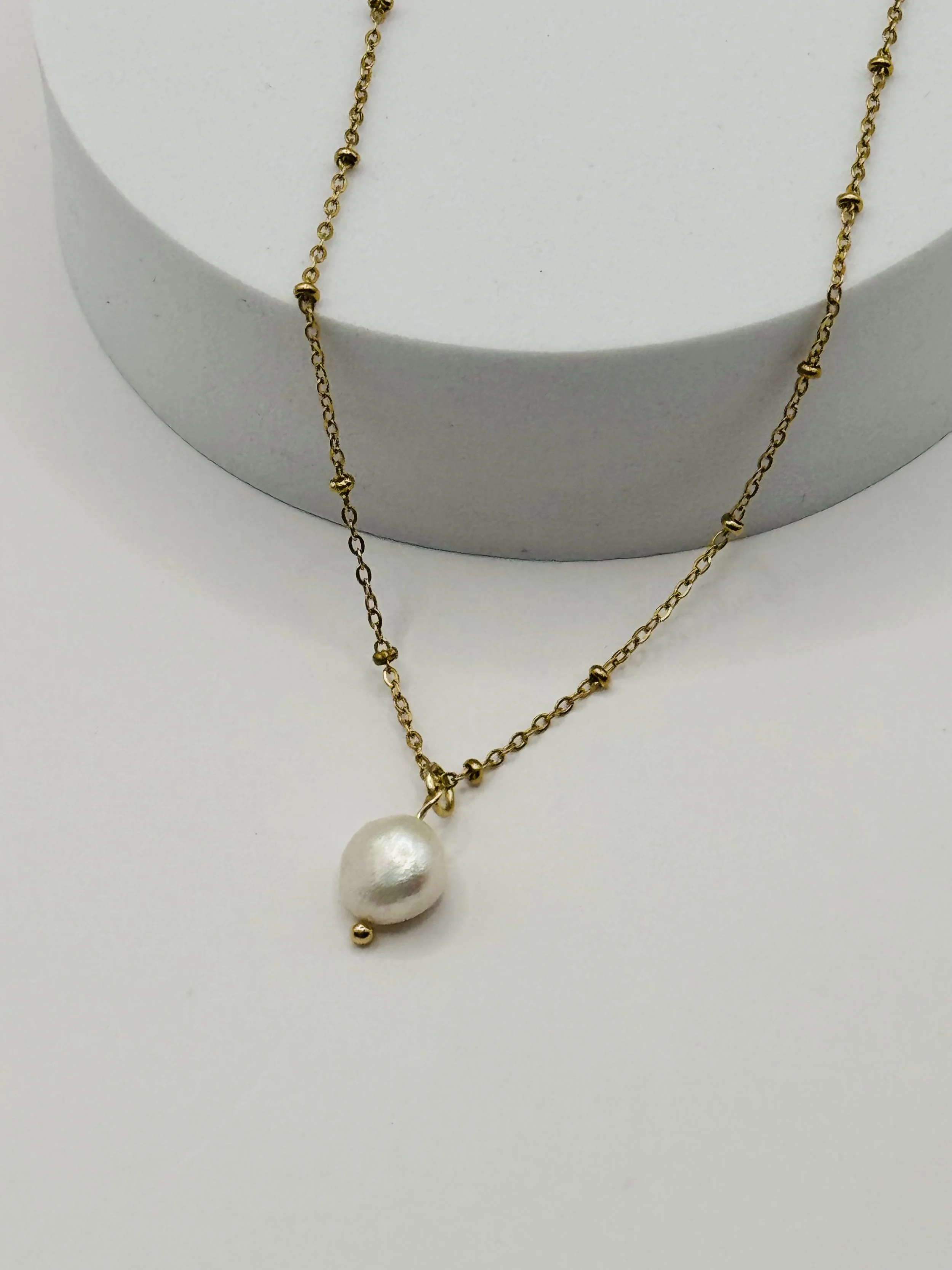 Gold necklace with a large white pearl pendant and small gold beads, displayed on a white surface with a grey cylindrical stand in the background. Available from Chain Rule Jewellery.