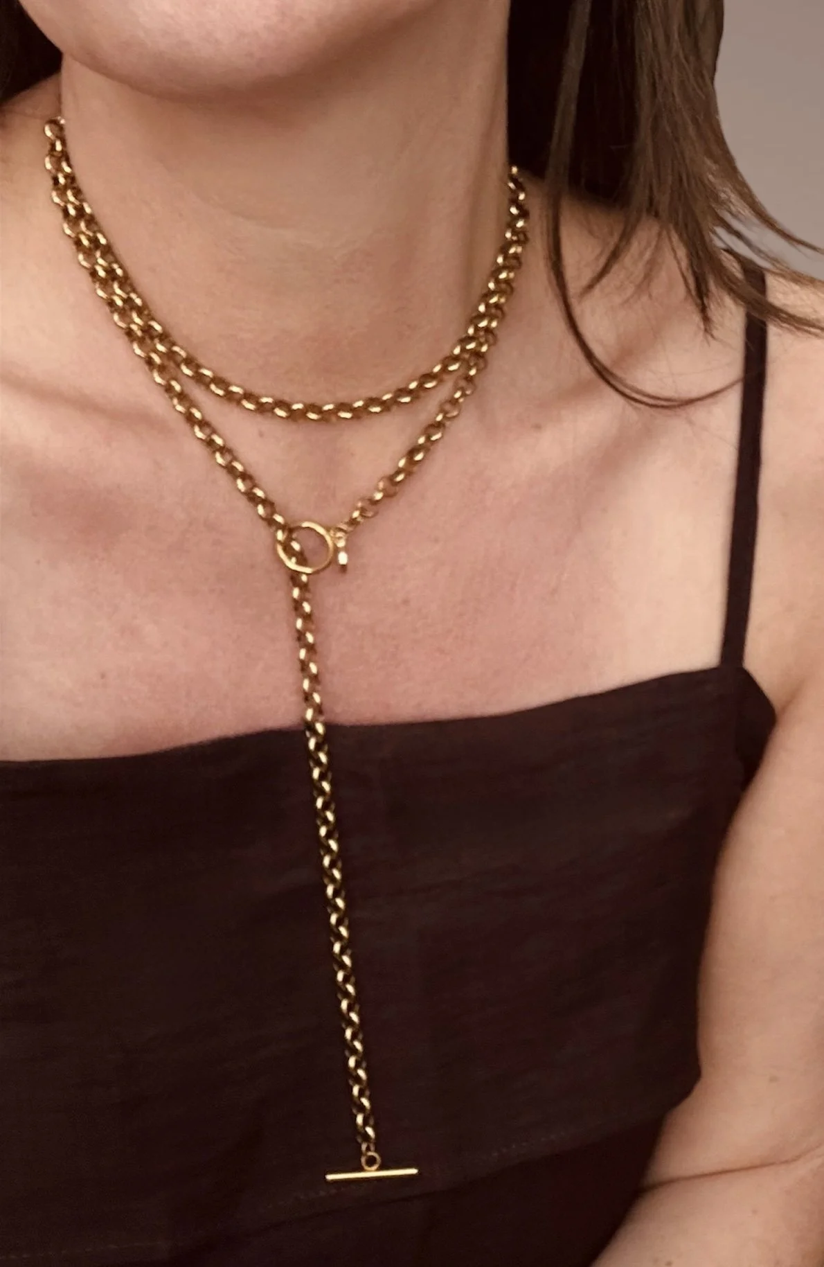 Close-up of a woman's neck wearing a layered gold chain wrap necklace, with a bar pendant, against a brown strap top.
