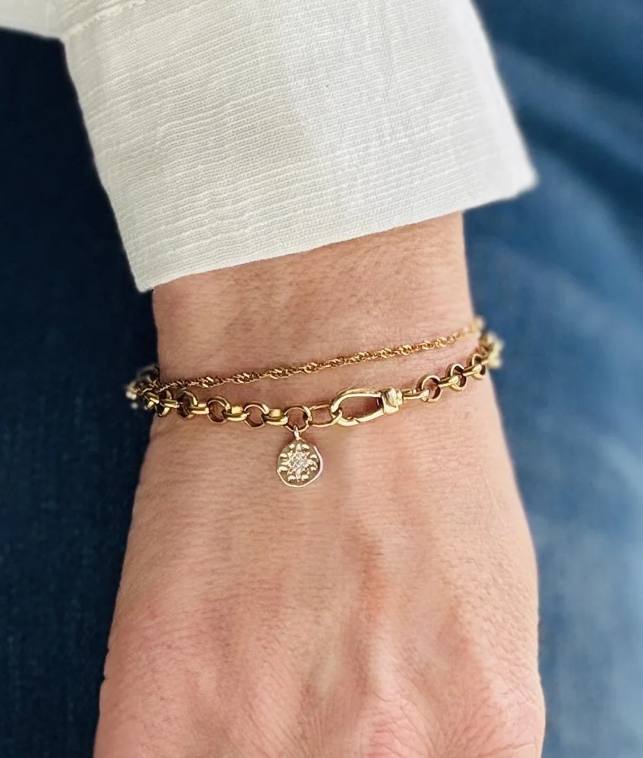 Gold bracelet with a small round charm hanging from it on a person's wrist, with a cream-colored sleeve and a dark blue fabric in the background.