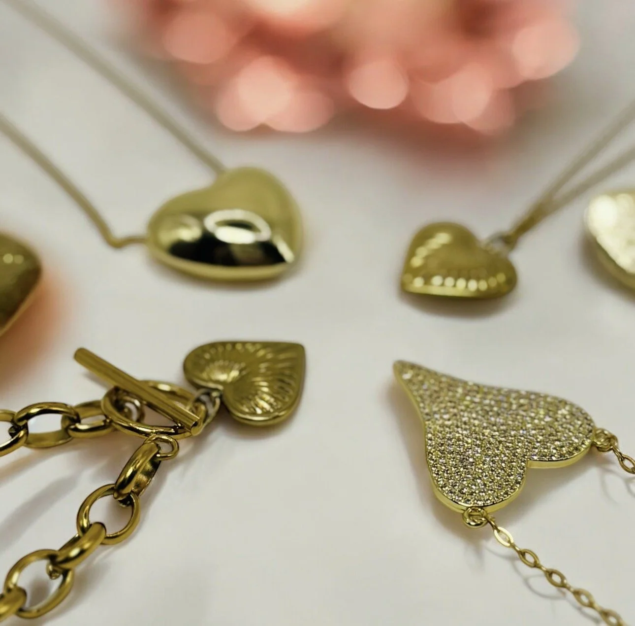 Gold heart-shaped jewelry pieces with chains laid on a white surface, with pink blurred background.