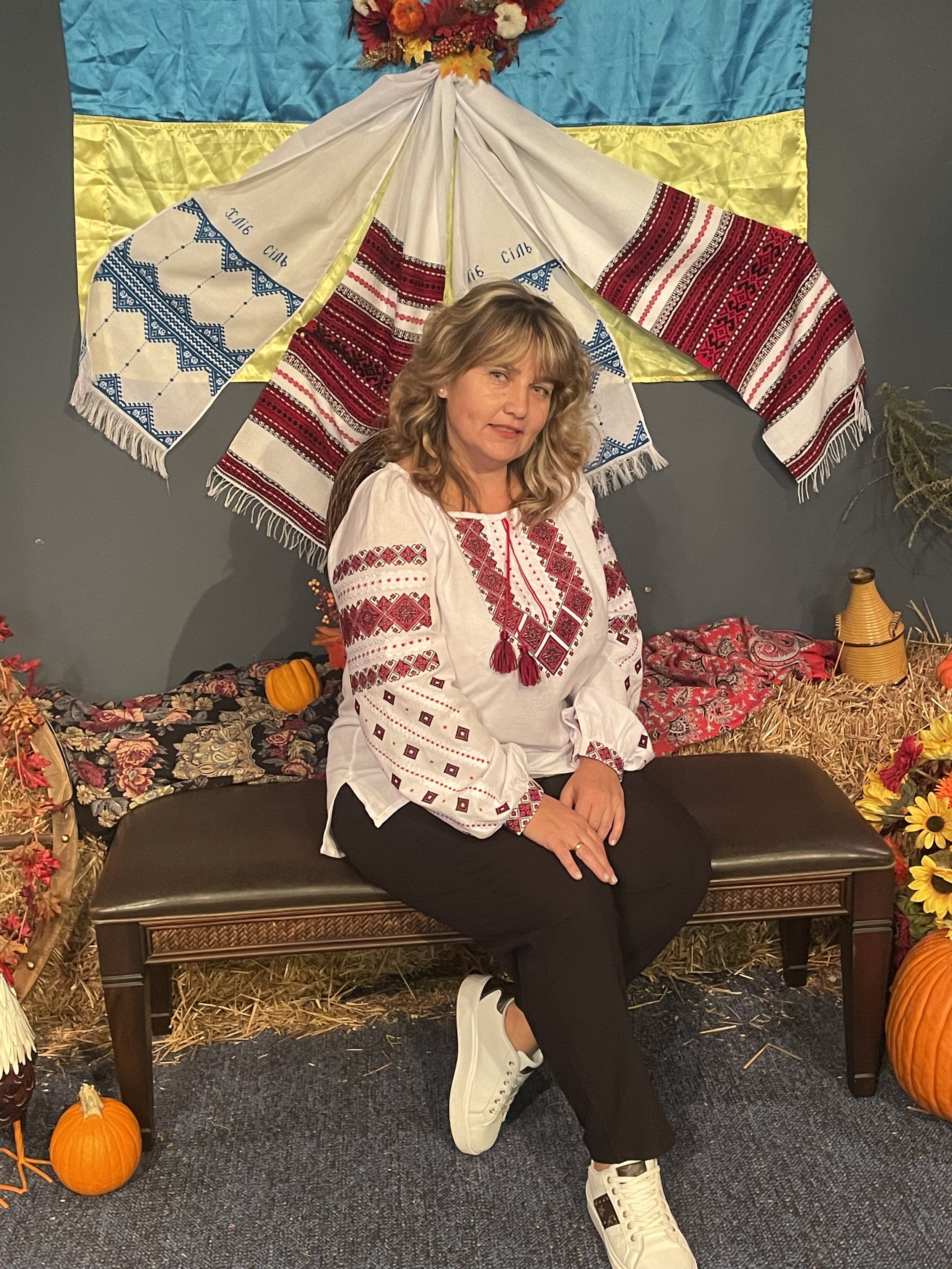 Woman wearing a white embroidered blouse sitting on a black bench in front of a colorful fall-themed backdrop with pumpkins, sunflowers, and hay, with a blue, yellow, and embroidered fabric hanging behind her.