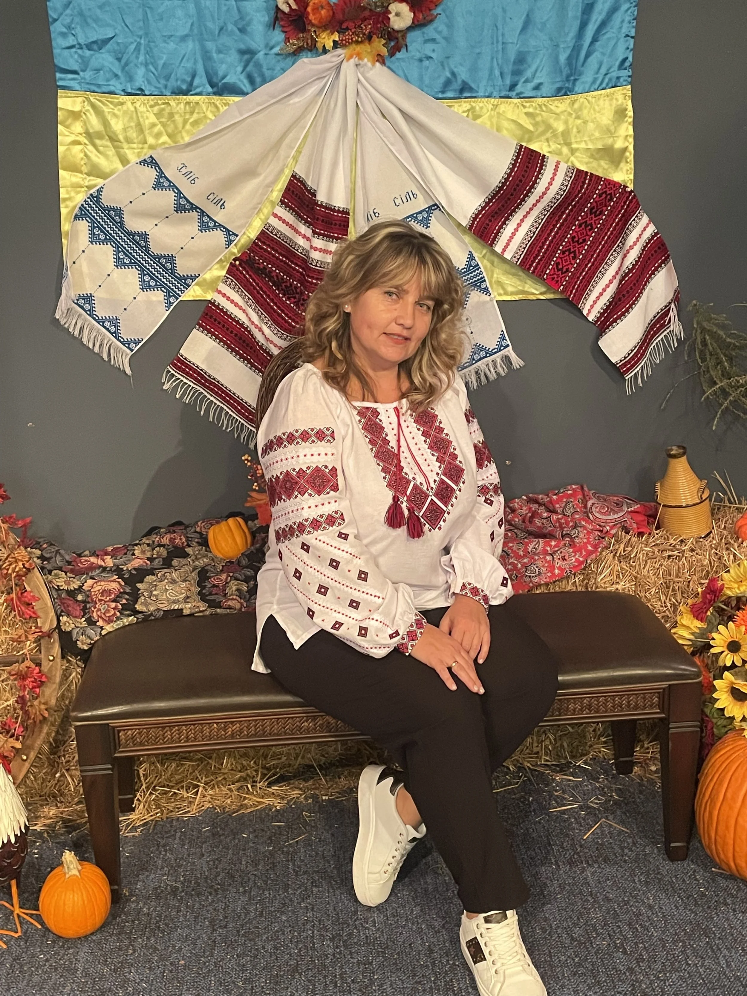 A woman sitting on a black bench with one leg crossed over the other, wearing a traditional embroidered blouse, black pants, and white sneakers. She is in front of a backdrop featuring a blue and yellow flag, hanging fabric, and autumn decorations including pumpkins, flowers, and hay bails.
