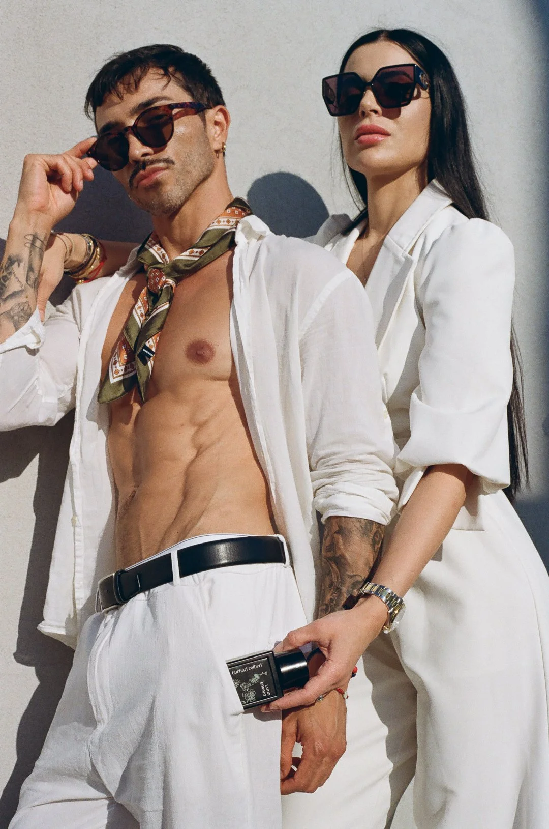 A man and woman posing together against a wall, both wearing stylish sunglasses. The man is shirtless with tattoos and a scarf, holding a perfume bottle. The woman is dressed in a white blazer and pants, with long black hair and a watch.