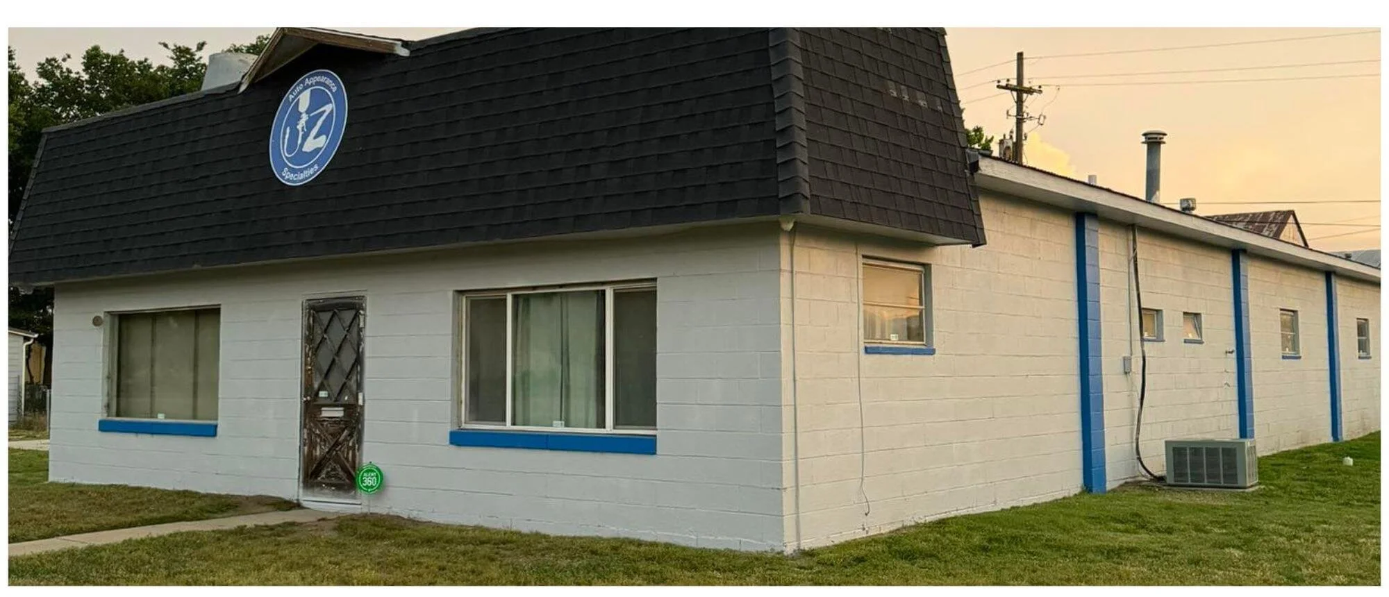 A single-story white brick building with blue trim and a black shingled roof, featuring a supermarket logo on the roof, windows with blue trim, and an air conditioning unit outside. The building is situated on a grassy lawn with a sidewalk in front.