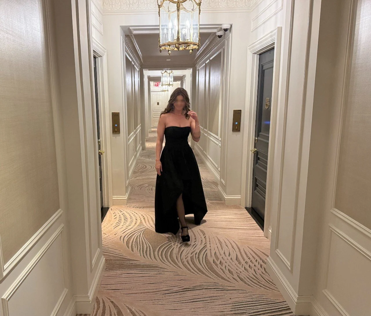 A woman in a black strapless gown taking a mirror selfie in an upscale hotel hallway with elegant wall paneling, a chandelier, and elevator doors.