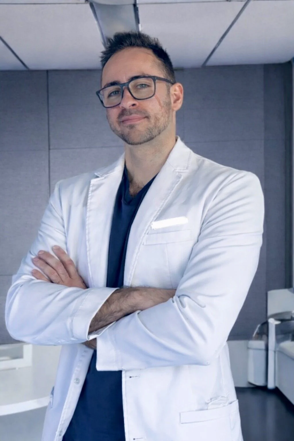 A male doctor with glasses, wearing a white lab coat and a dark shirt, standing with arms crossed in a clinical setting with gray walls.