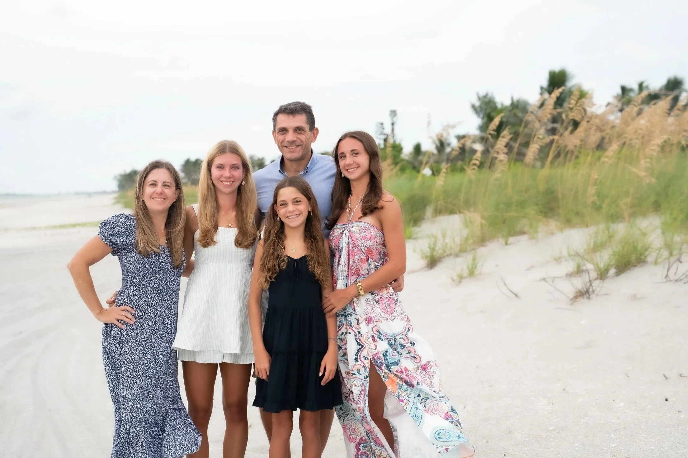 A family of six standing on a beach with sand dunes and greenery in the background, smiling at the camera.