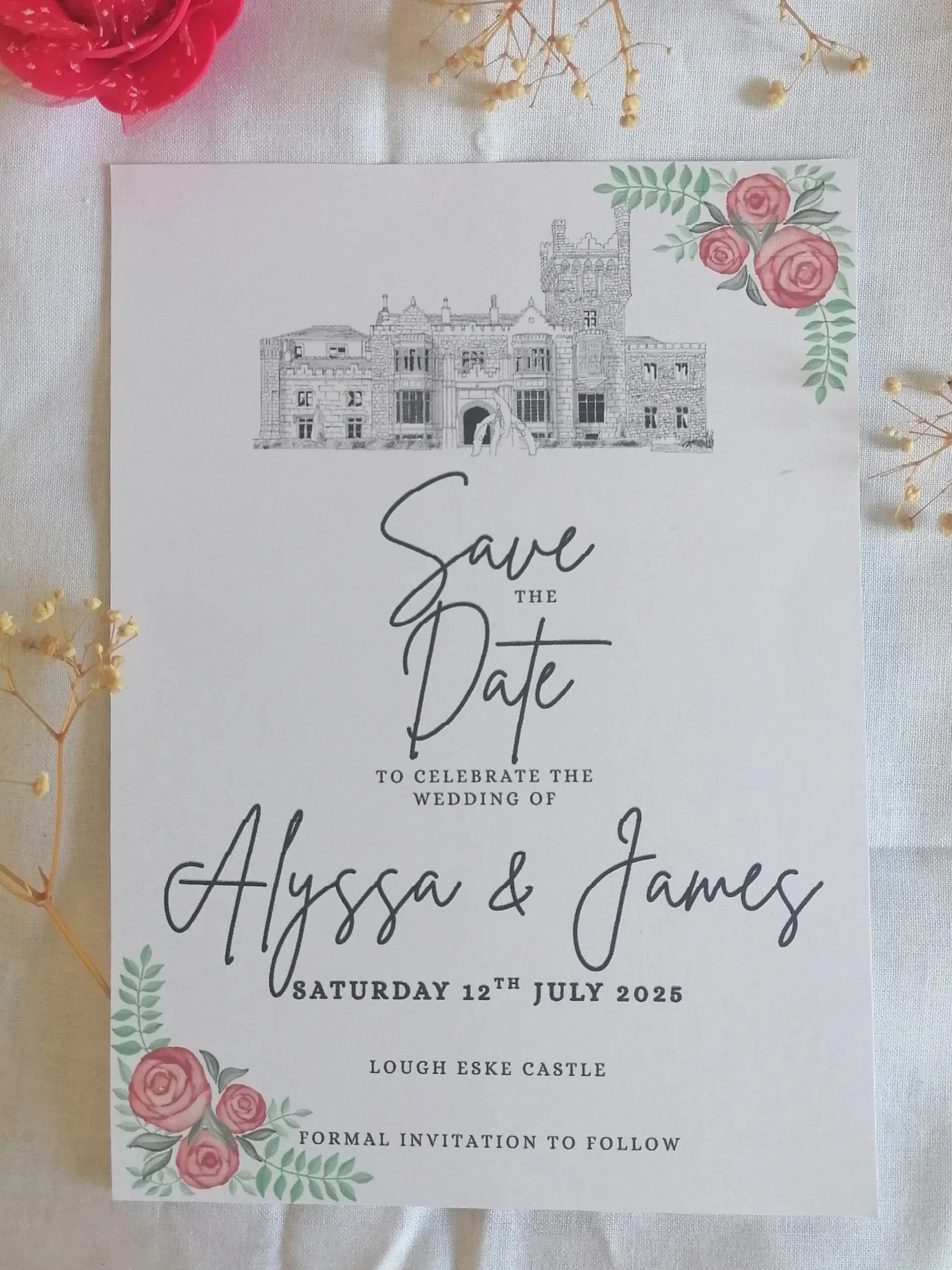 A wedding save-the-date card featuring a castle illustration at the top, floral decorations with pink roses at the top right and bottom left corners, and text announcing the wedding of Alyssa and James on Saturday, July 12, 2025, at Lough Eske Castle. The card is placed on a white surface with small decorative flowers around it.