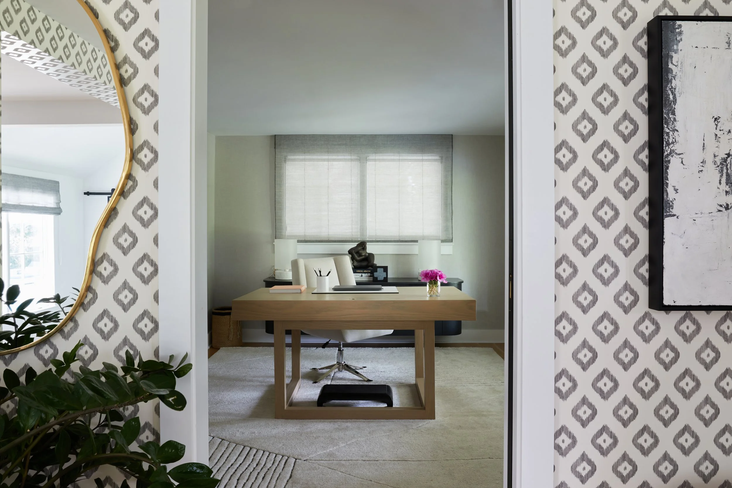 A home office with a wooden desk, white chair, and decorative items in front of two windows with sheer gray curtains. The room has light-colored walls and a light-colored carpet. A sculpture and a small plant with pink flowers are on the desk.