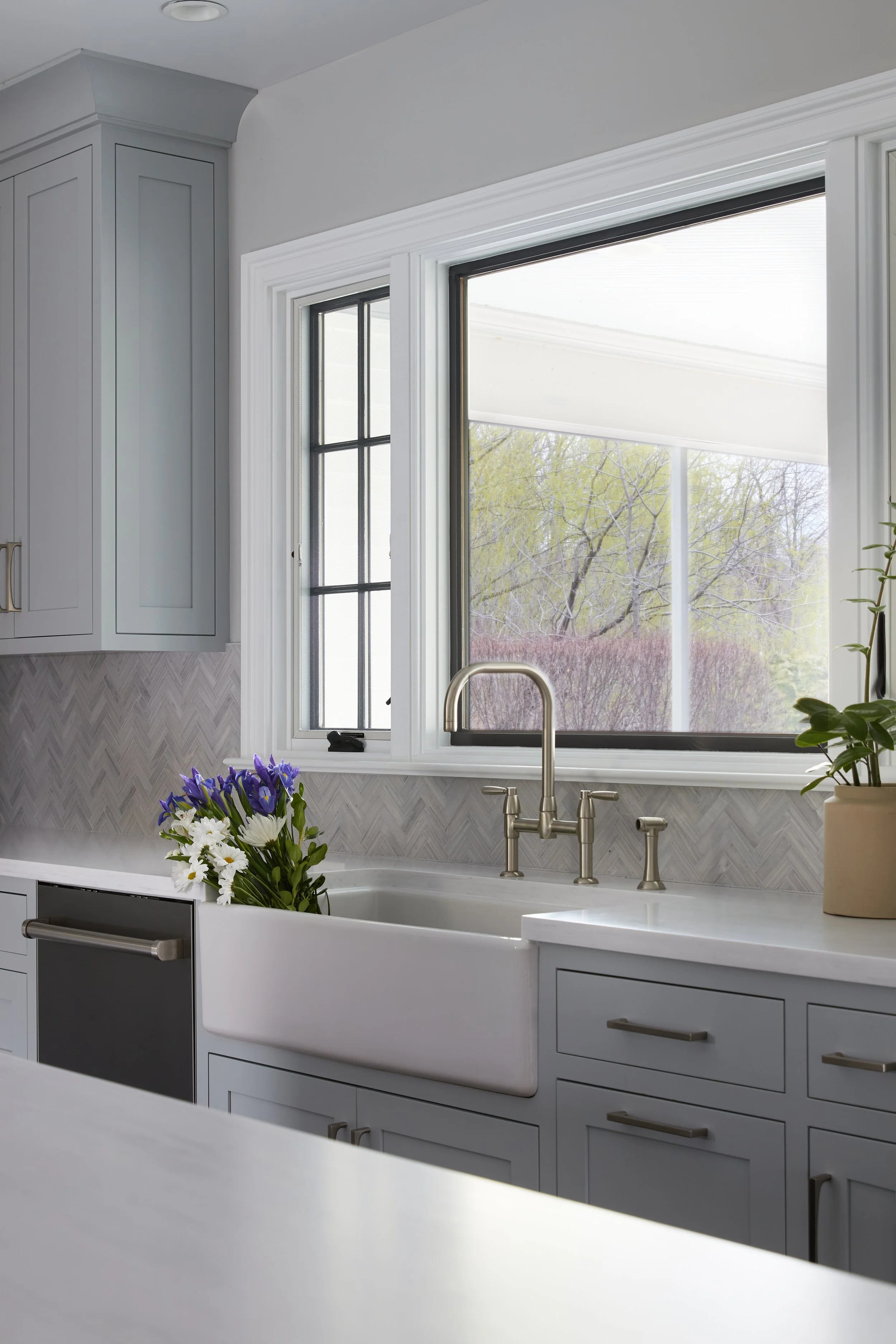 A bright kitchen with a farmhouse sink, a floral bouquet inside the sink, and a large window showing an outdoor scene with trees.