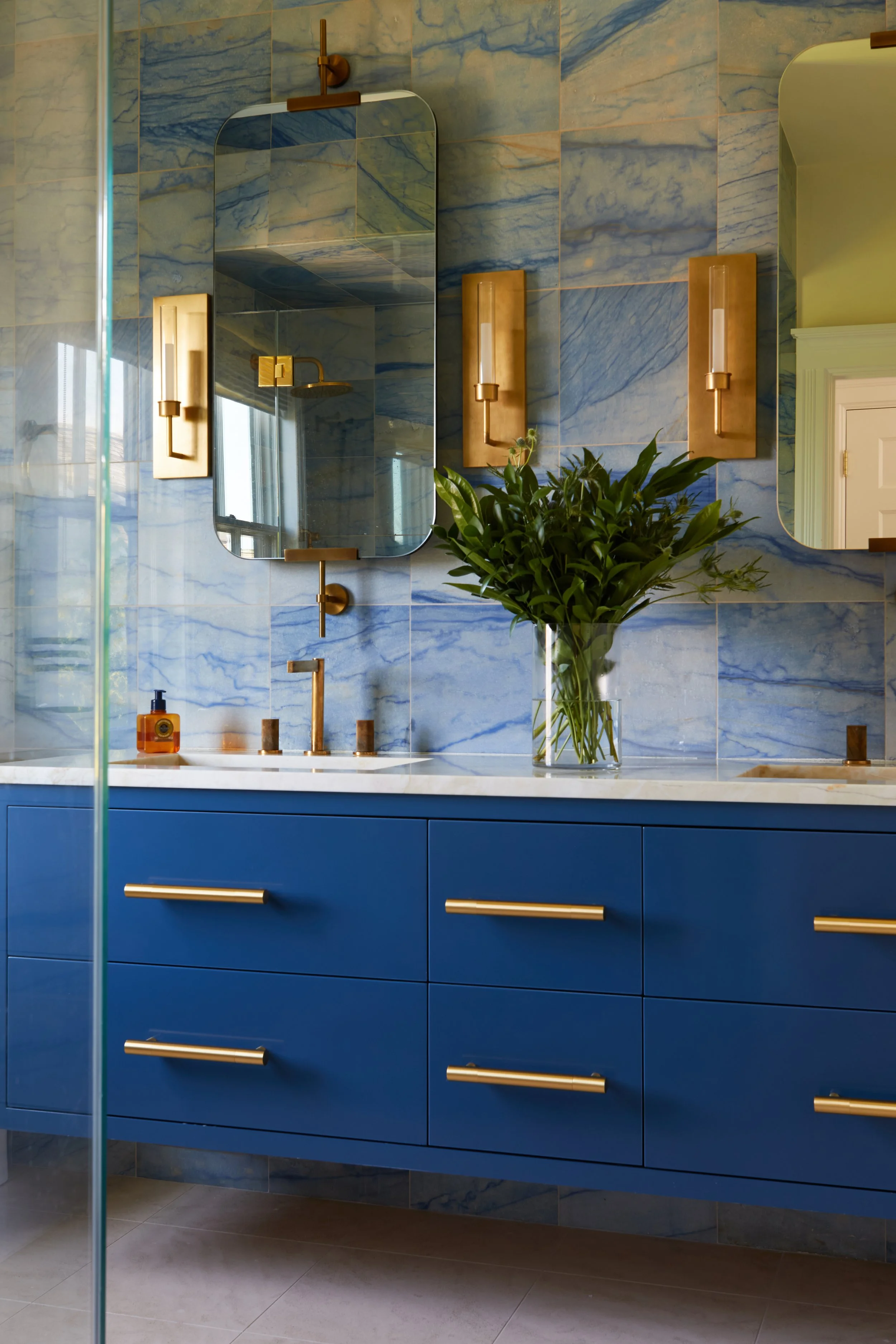 A modern bathroom with a blue vanity and gold handles, a white marble countertop, a large mirror, a vase with green foliage, a bottle of soap, and gold wall-sconces against a blue marble wall.