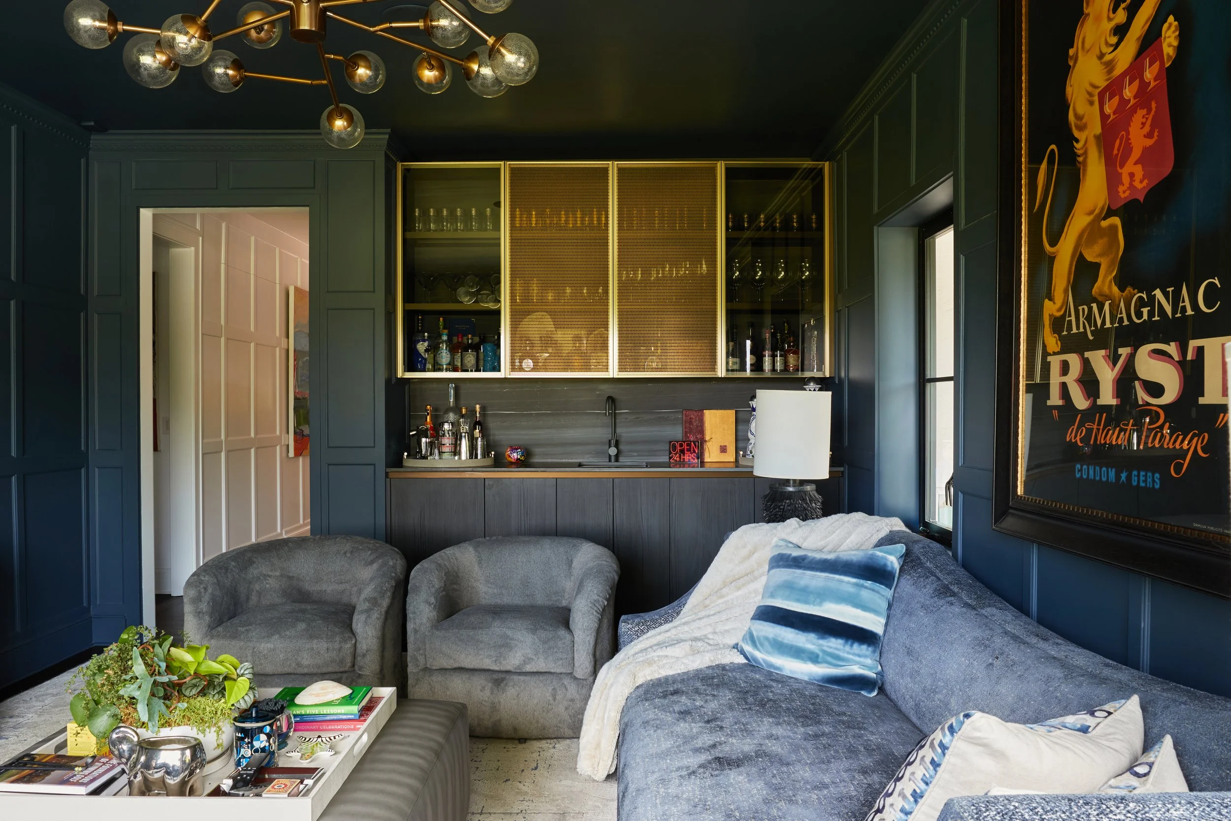 Living room with blue wall panels, a black bar with gold trim, bar supplies, two gray armchairs, a gray sofa with pillows, a coffee table with decor, and a large framed poster with a lion and text.