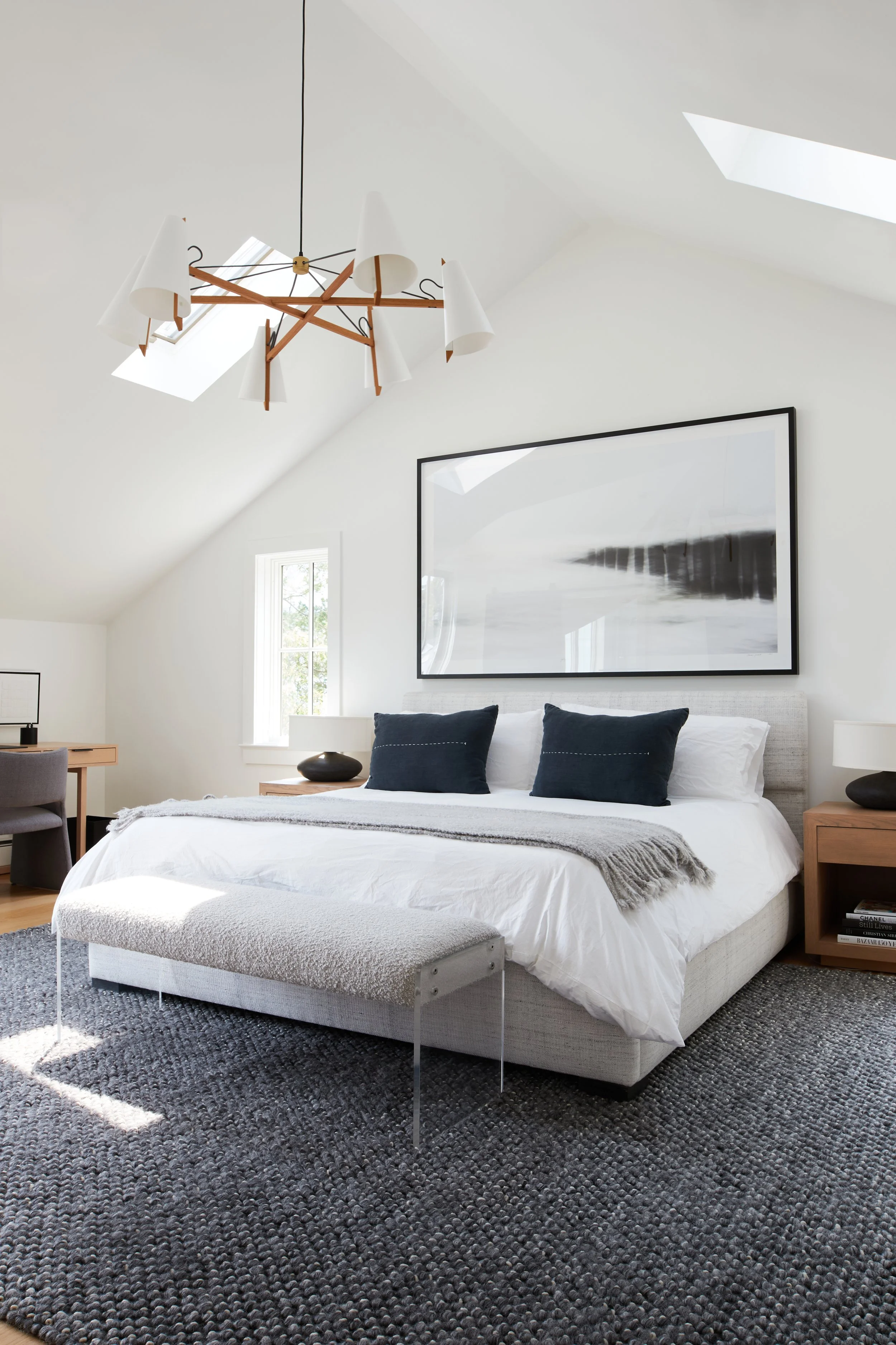 Modern bedroom with a white bed, navy pillows, and a large black-and-white artwork above, illuminated by natural light from skylights and a window.