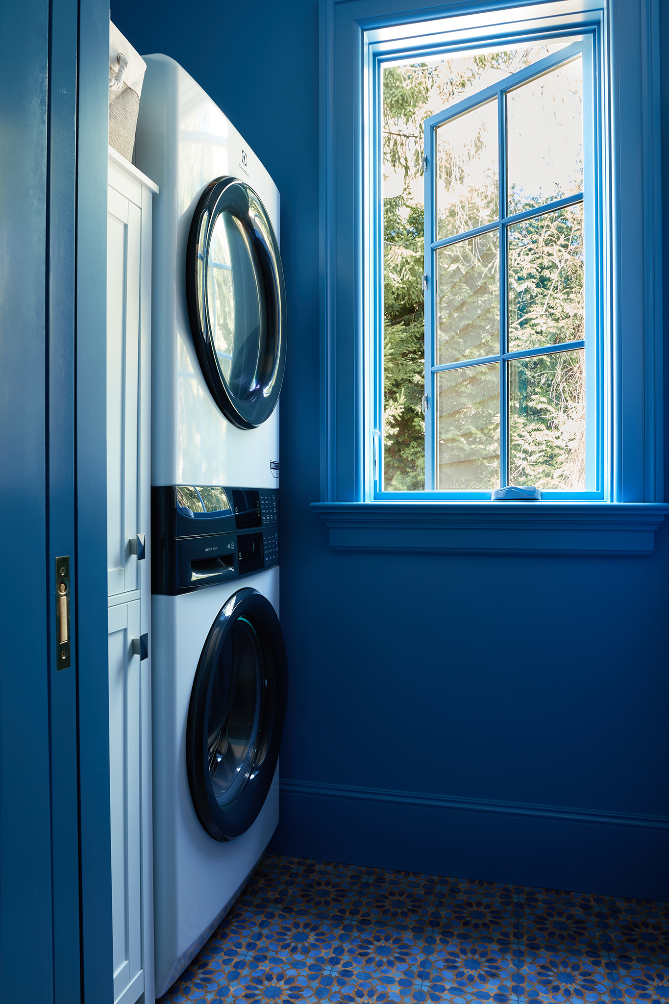 A laundry room with a stacked washing machine and dryer next to a window showing trees outside, and a blue wall and cabinetry.