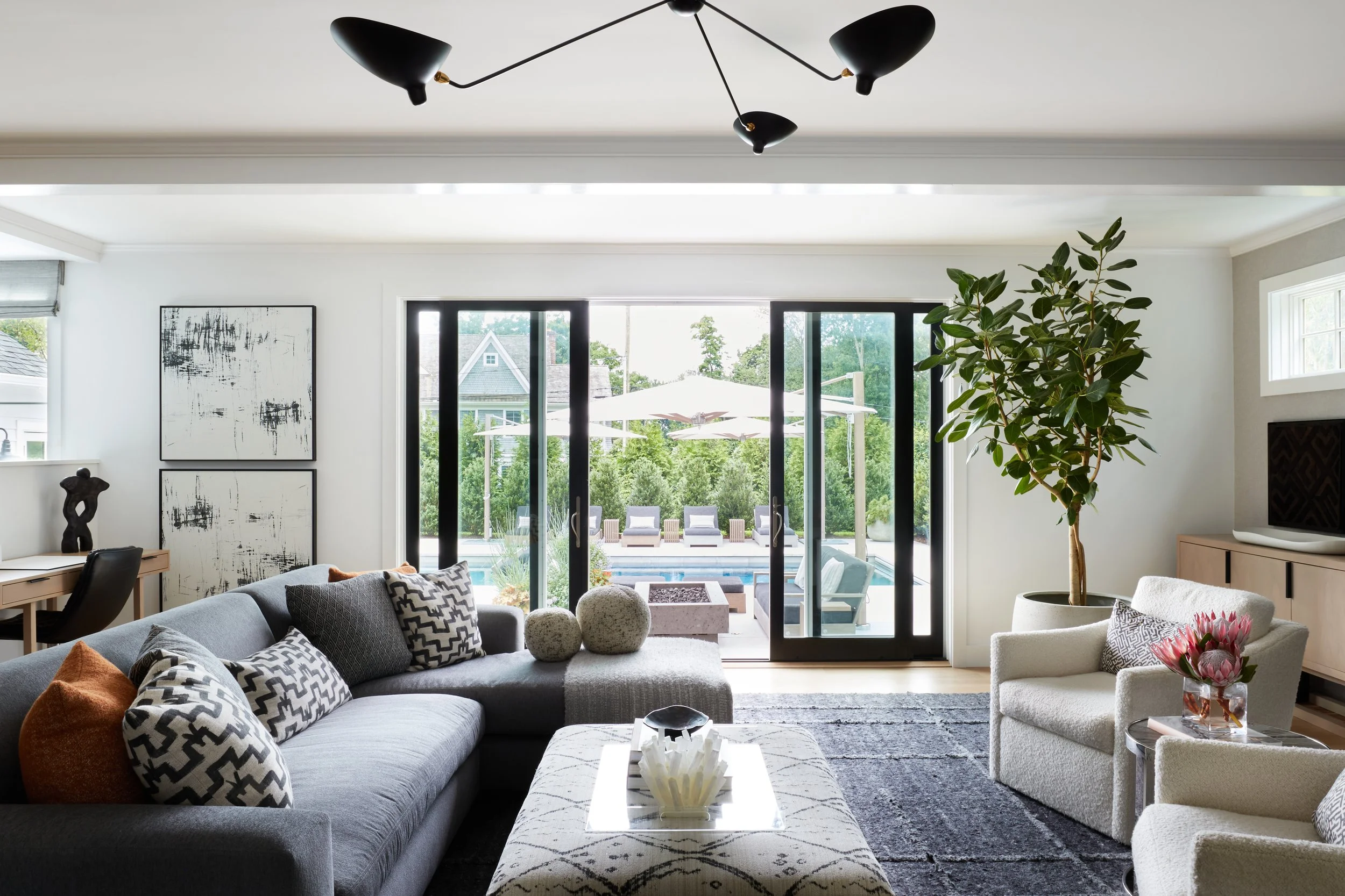 Modern living room with gray sectional sofa, white armchairs, abstract black-and-white artwork, large potted plant, glass sliding doors to outdoor patio with pool and seating area.