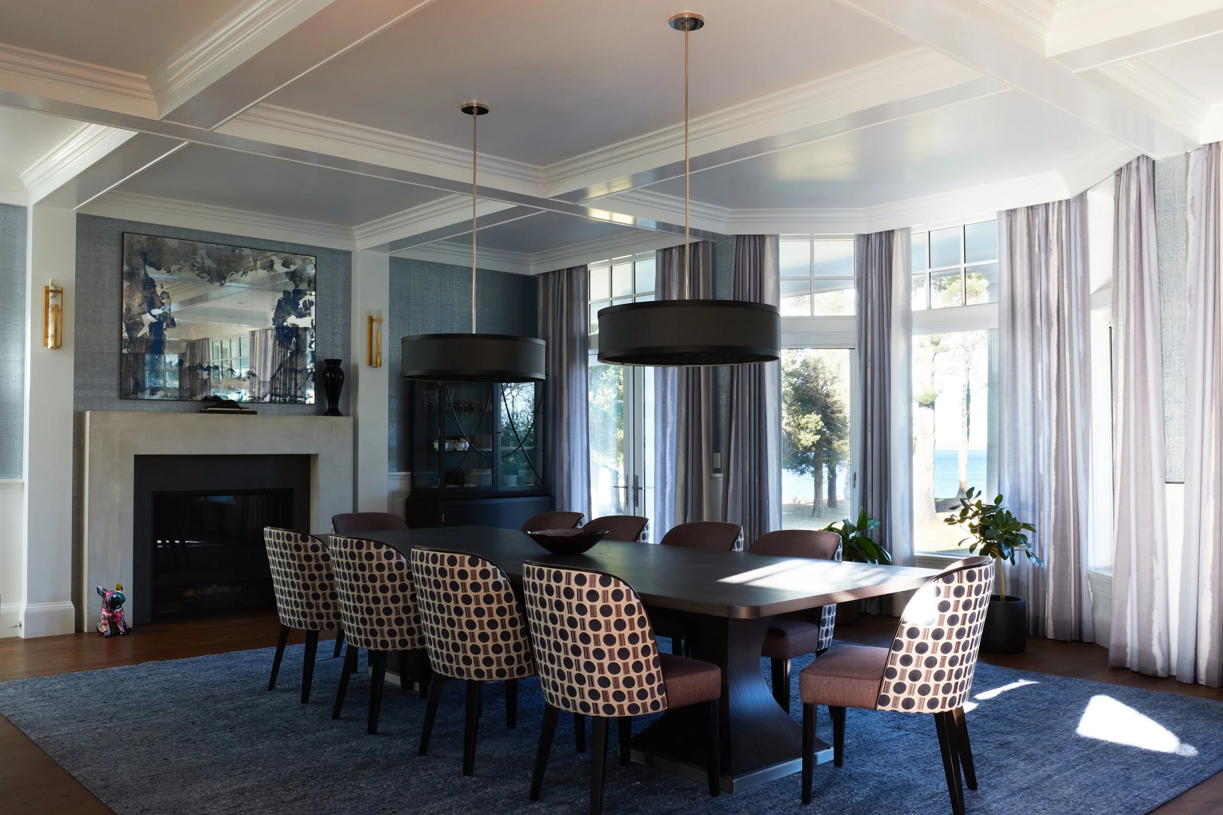 Bright dining room with large windows, round black pendant lights, a rectangular wooden table with patterned chairs, a fireplace, a mirror above the fireplace, and decorative vases, with sunlight streaming in through curtains and outside view of tree