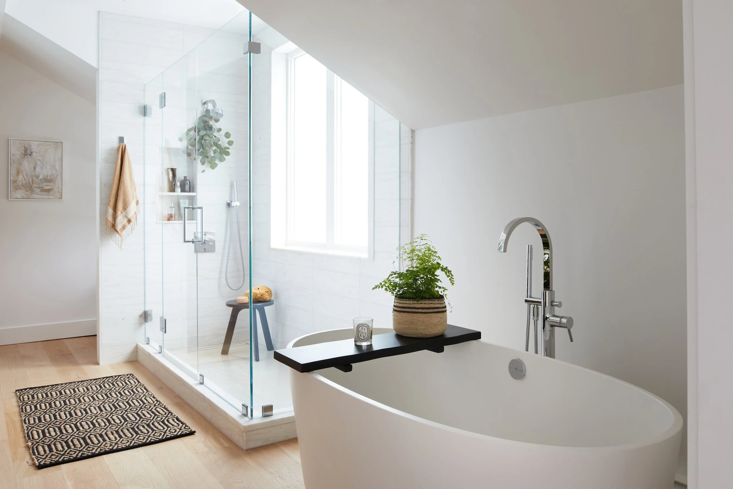 Modern bathroom with a glass-enclosed shower, freestanding bathtub, wooden floor, potted plant, and a window letting in natural light.