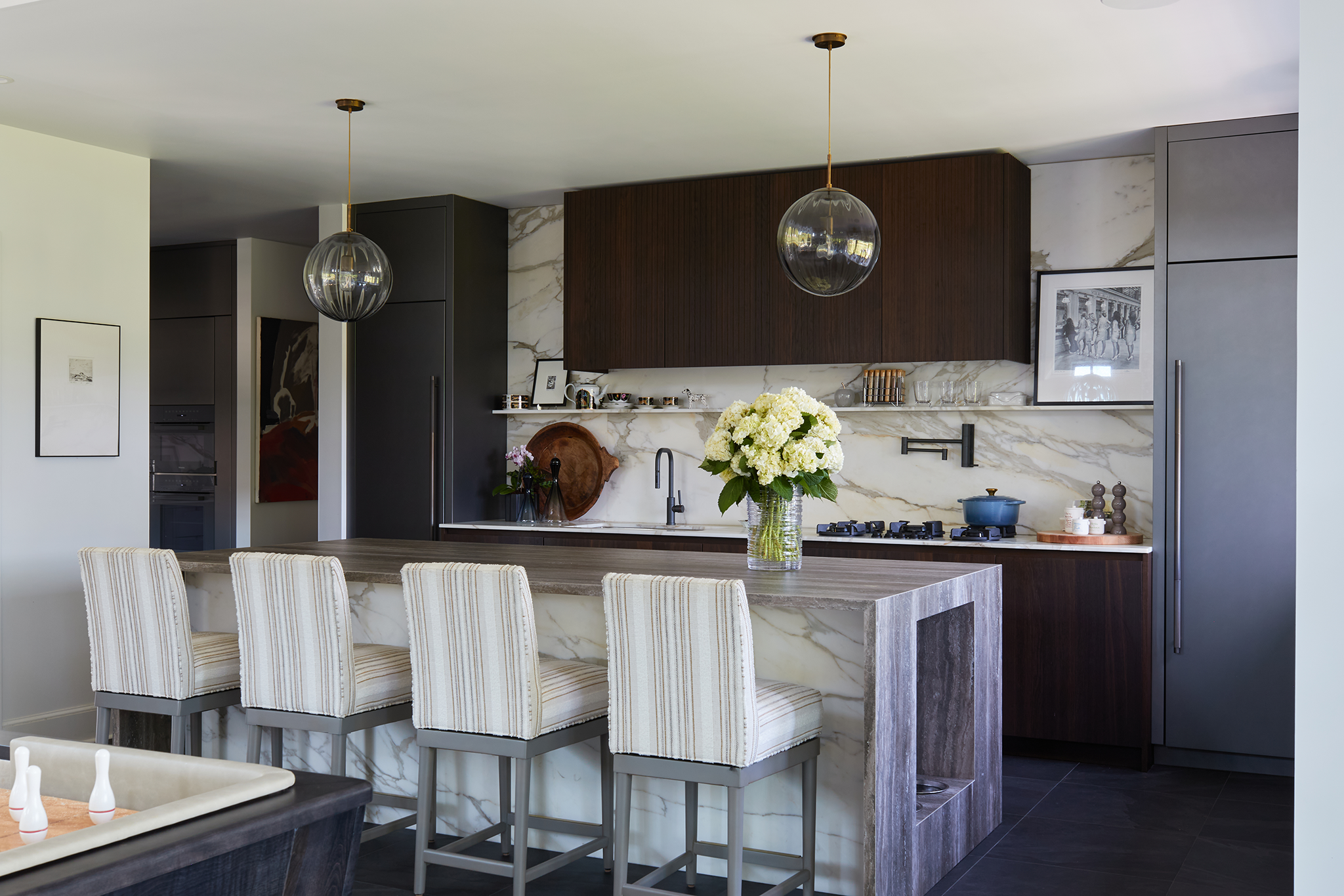 Modern kitchen with marble countertops, dark wood cabinets, a large vase with white hydrangeas, and barstools at a marble island.