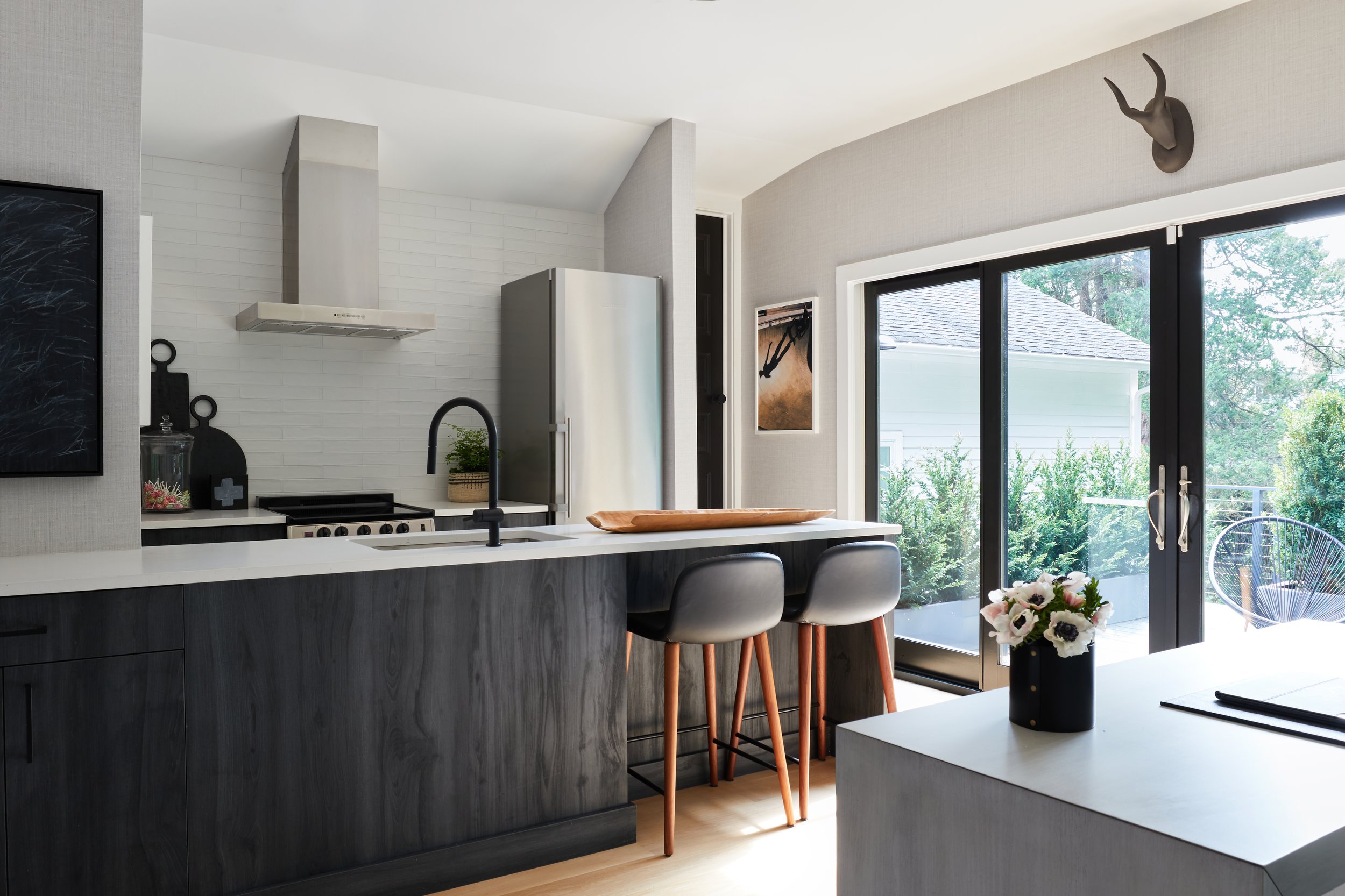 Modern kitchen with black and white color scheme, stainless steel refrigerator, black countertop, white walls, and large sliding glass door leading to outdoor patio with greenery.
