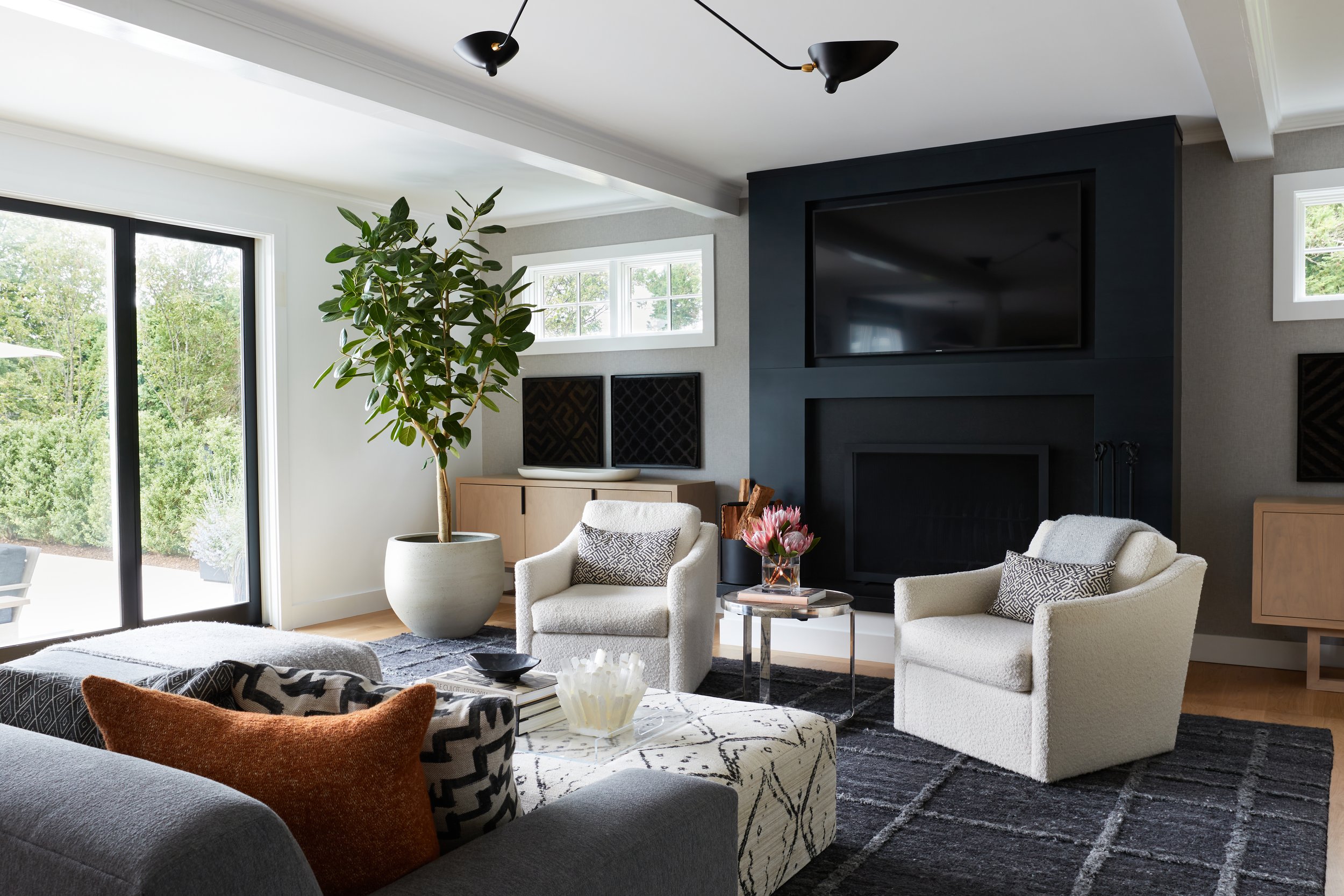 Modern living room with white armchairs, a sofa with patterned pillows, a glass coffee table, a large potted plant, a black television above a fireplace, a wooden sideboard, and large windows showing greenery outside.