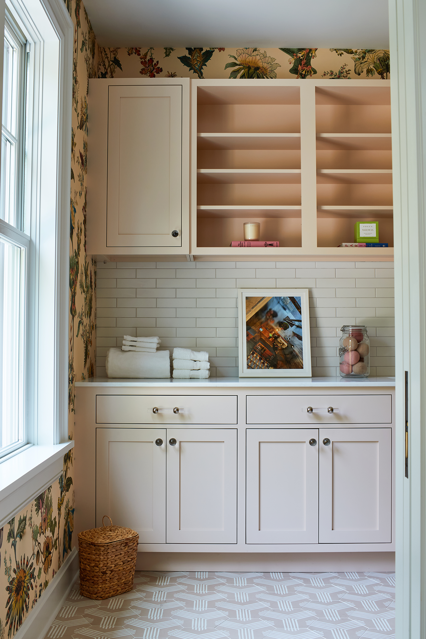 A laundry room with floral wallpaper, white cabinets, open shelves, a white countertop, stacks of white towels, a framed picture, a glass jar with pink and beige bath bombs, and a wicker basket on the floor.