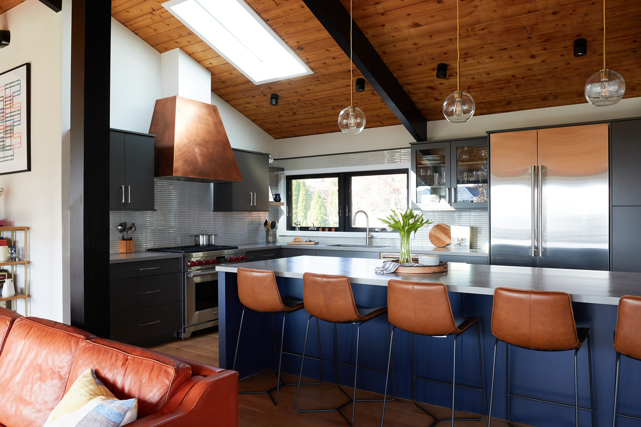 Modern kitchen with a wooden ceiling, skylight, and black and gray cabinetry. A white countertop island with four brown leather barstools, glass pendant lights, and a vase of white flowers.