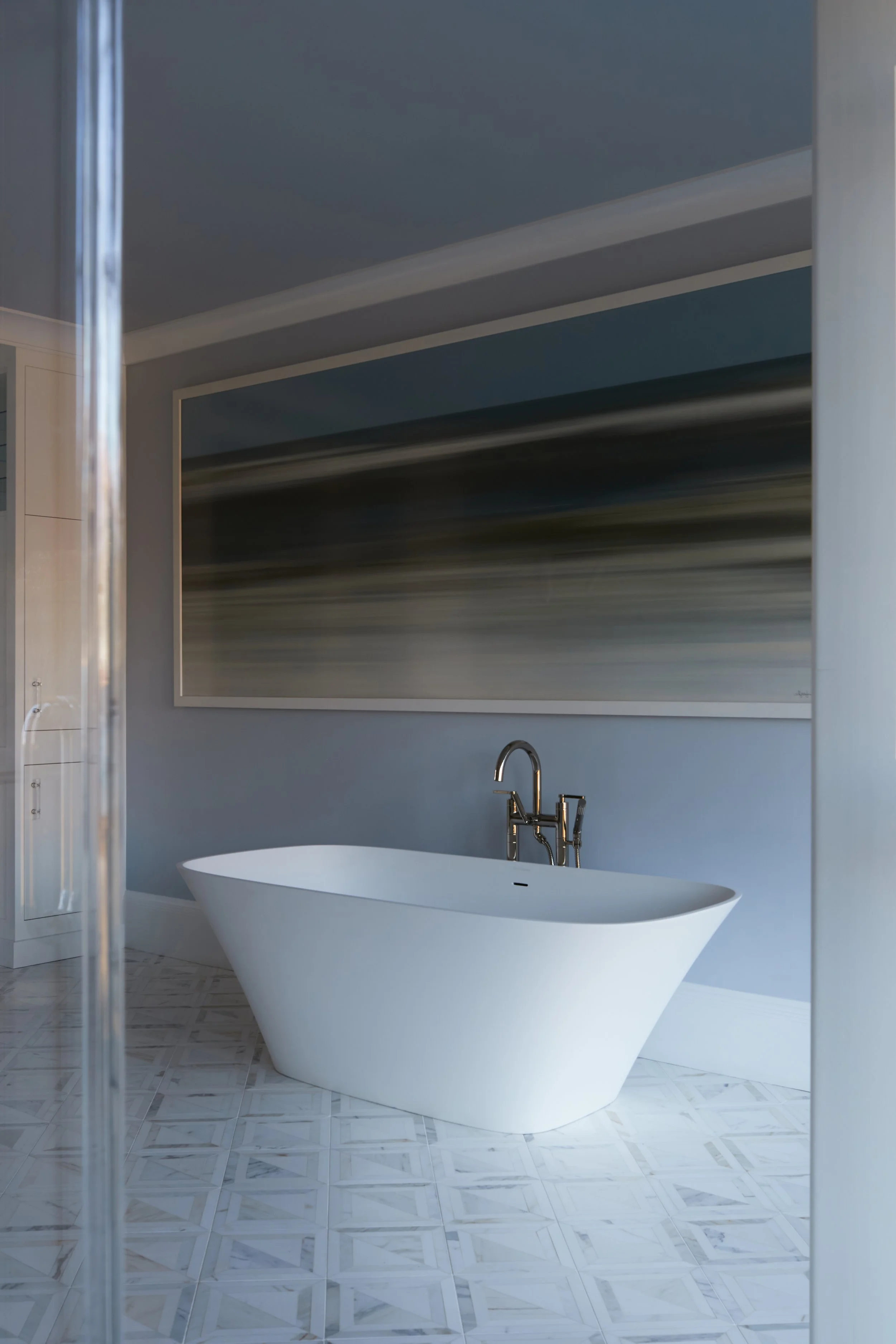 Modern bathroom with a white freestanding bathtub, a sleek chrome faucet, a large abstract painting on the wall, and patterned tile flooring.