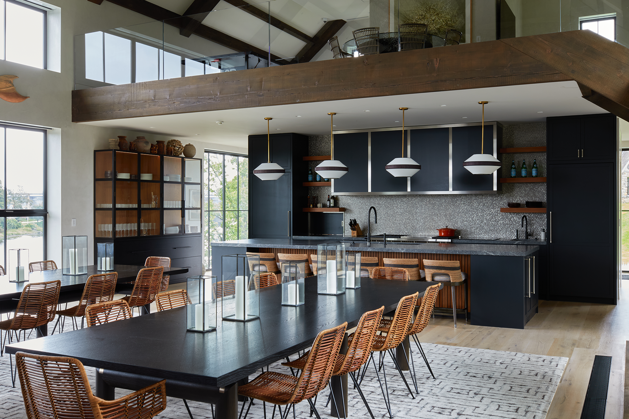 Modern kitchen and dining area with a large black dining table and wicker chairs, black kitchen cabinets, a gray marble countertop, wooden shelves, hanging pendant lights, and large windows providing natural light.