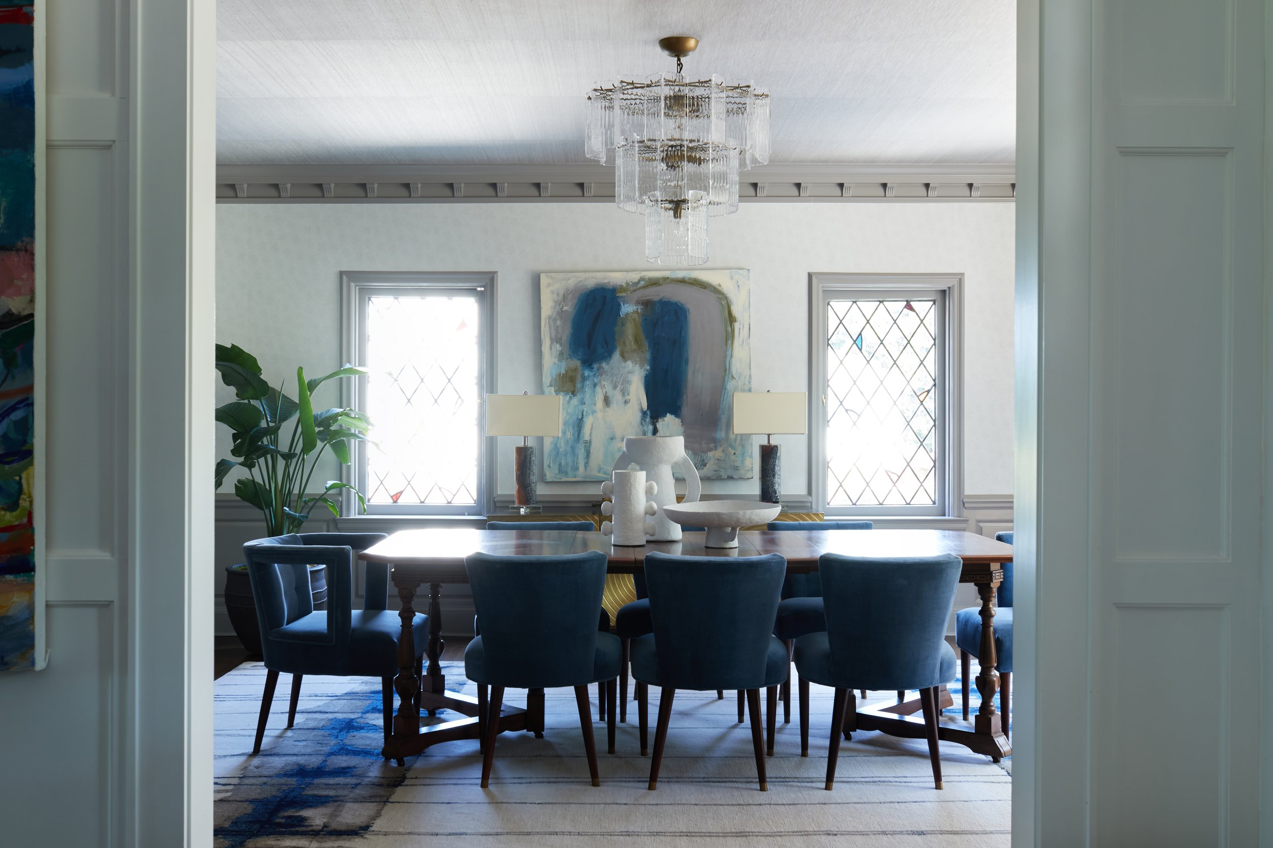 A dining room with a large wooden table surrounded by seven dark blue chairs. On the table are white ceramic vases and bowls. Two lamps and an abstract painting are on the wall behind the table. Two windows with diamond-shaped panes let in natural li