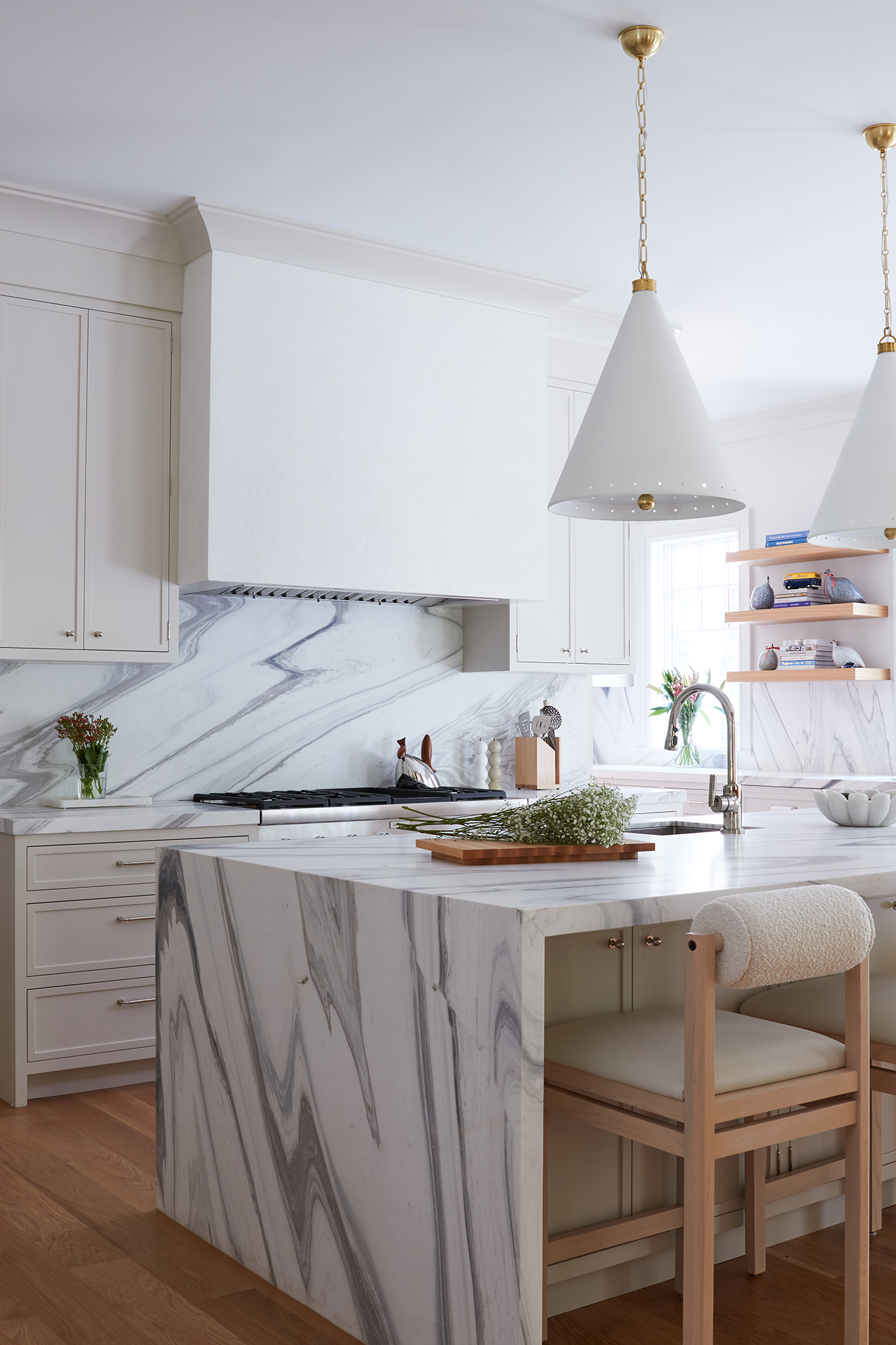 Modern kitchen with white cabinetry, marble backsplash, marble kitchen island, wooden stools with white cushions, pendant lights, and open shelves with books and decor.