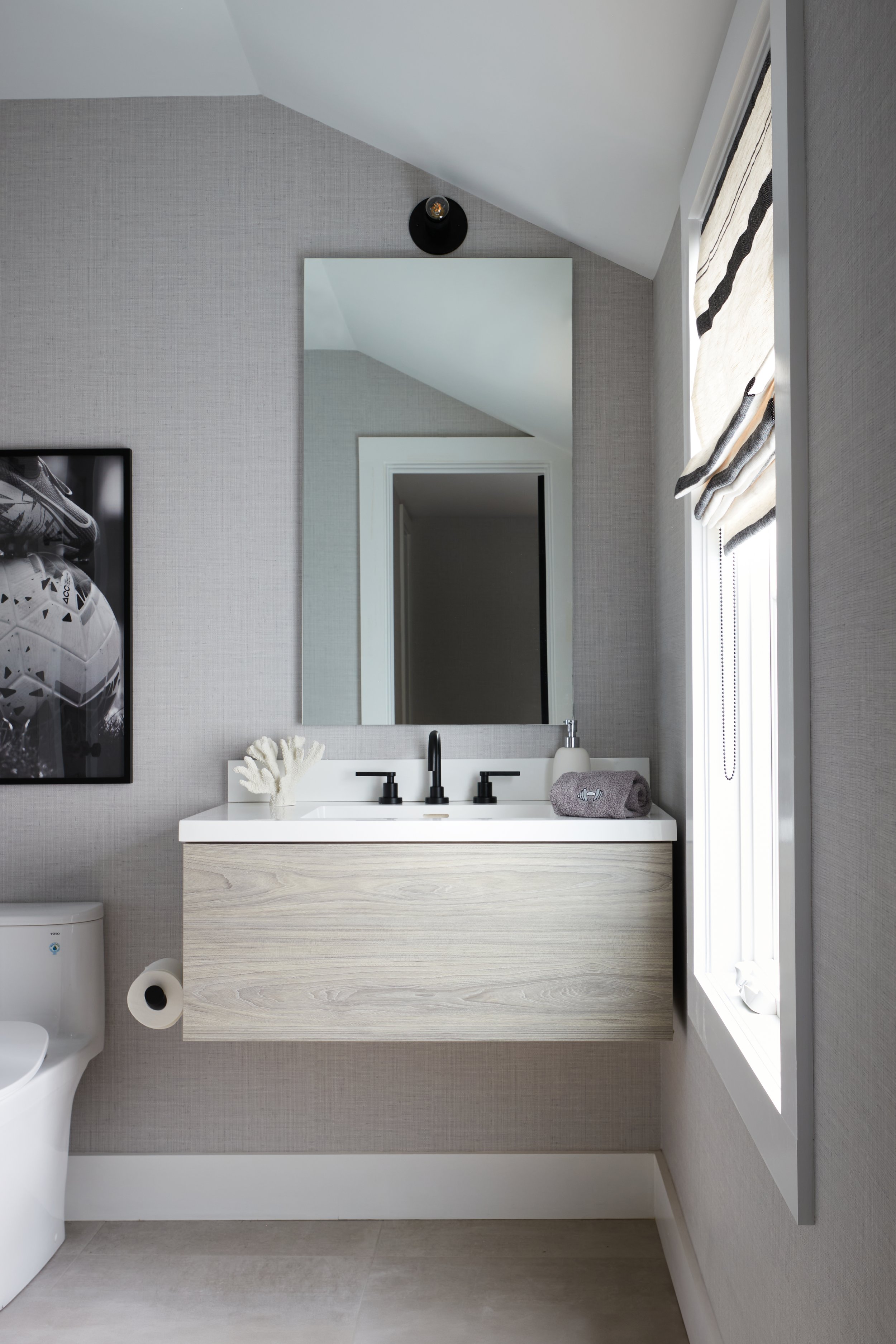 Modern bathroom featuring a floating wooden vanity with a white countertop, black faucet, mirror, window with a roman shade, and wall art of a sneaker.