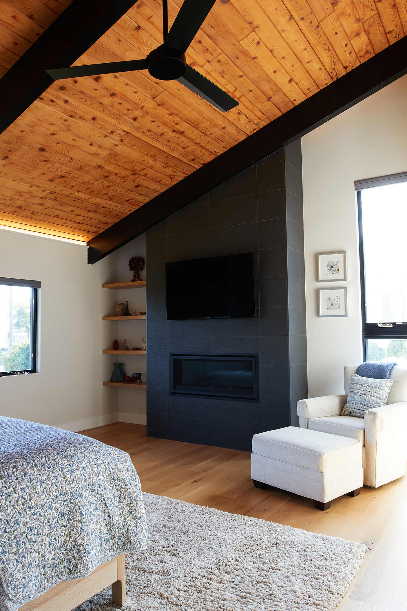 Living room with a vaulted wood ceiling, a black ceiling fan, two windows, a black tiled fireplace with a mounted TV, a white armchair with an ottoman, and built-in shelves with decorative items.
