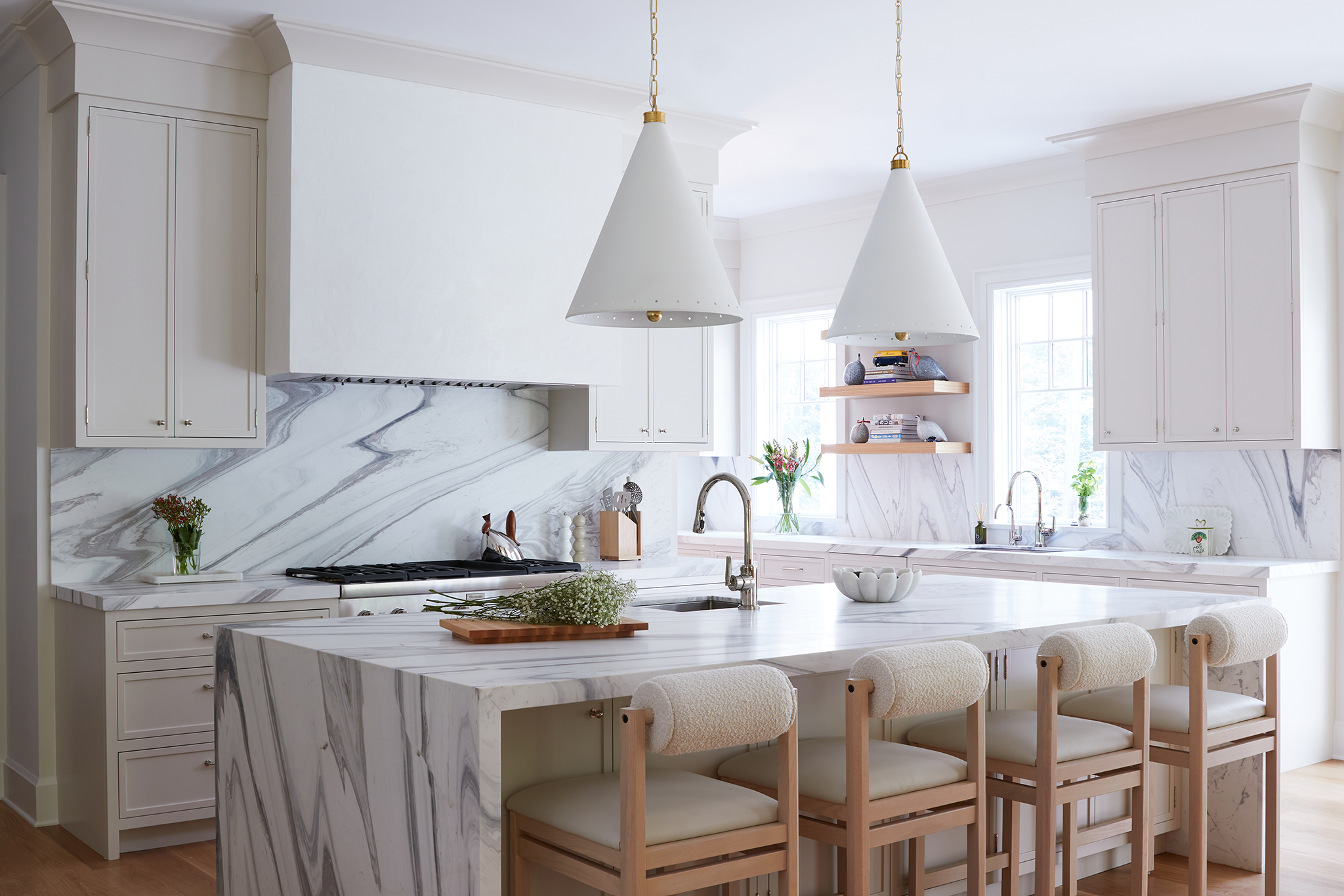 A bright, white kitchen with marble countertops and backsplash, three white bar stools with wooden legs at a marble island, white cabinets, two windows with natural light, and pendant lights above the island.
