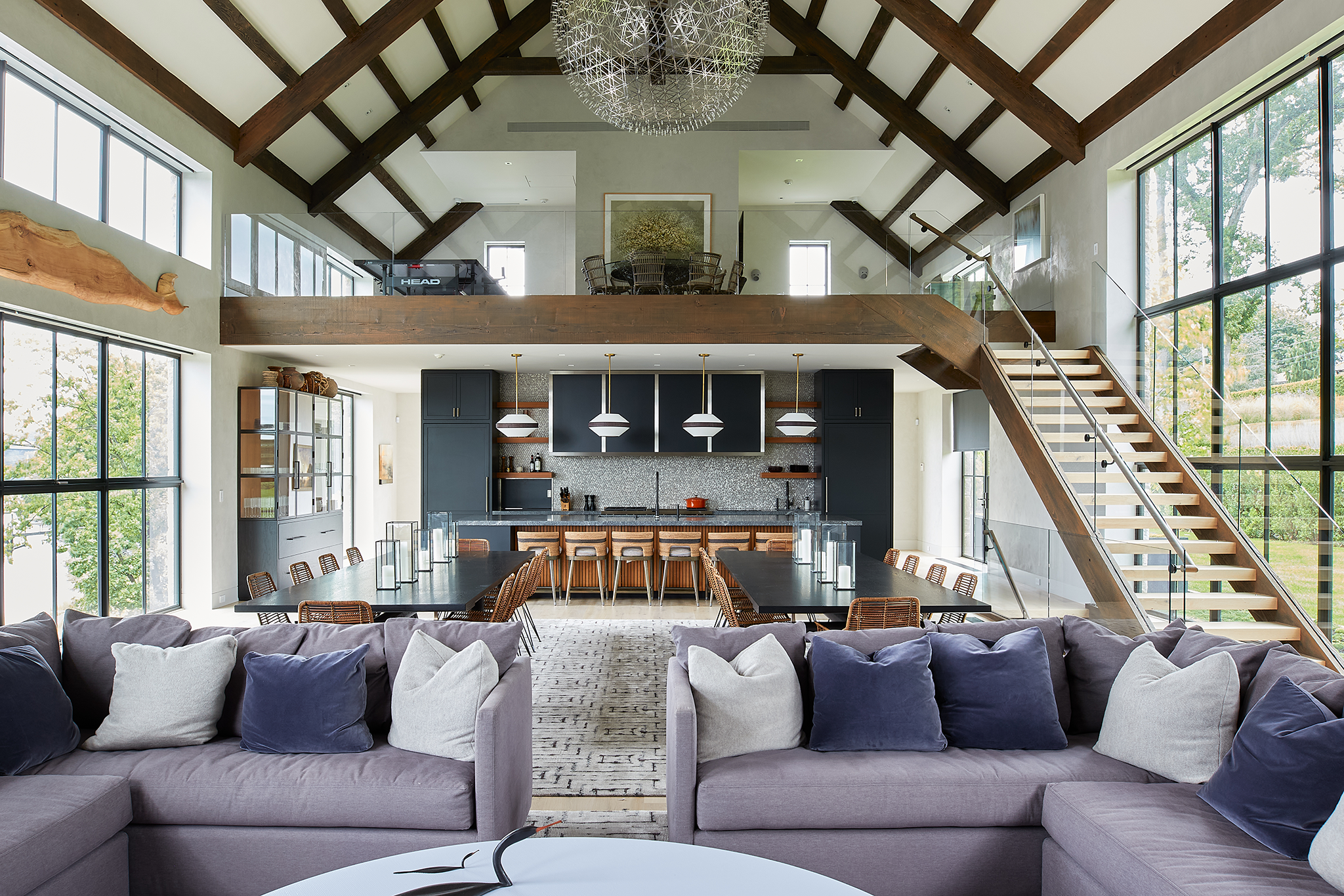 Open-concept living and dining area in a modern home, with large windows, a high vaulted ceiling with wooden beams, a staircase, and a kitchen in the background with navy cabinets and pendant lighting.