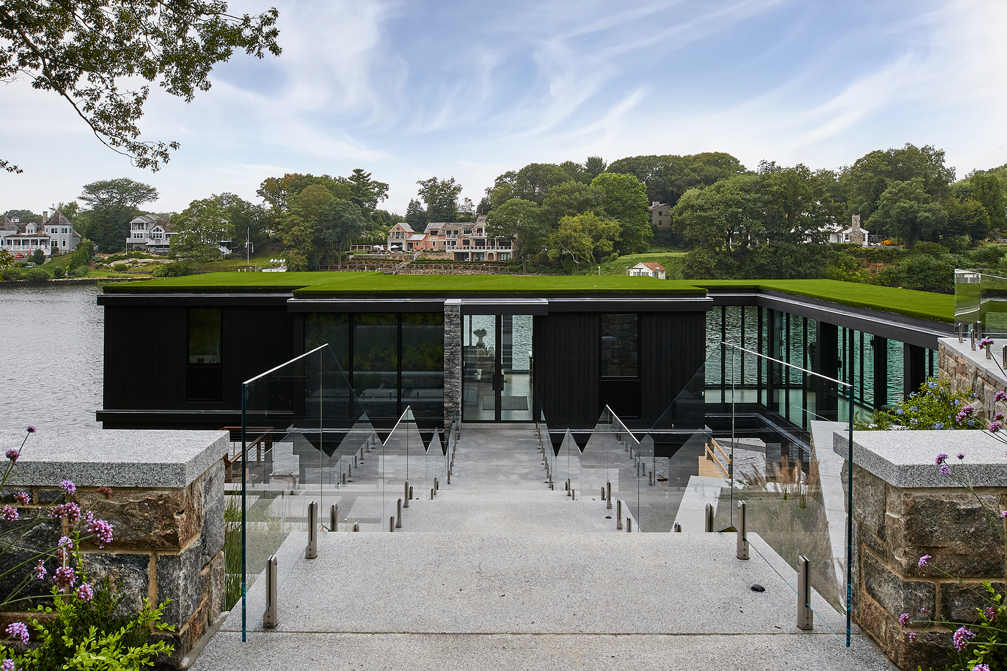 Modern house with black exterior and flat roof, built on a waterfront, with glass railing and steps leading down to the water. Background shows a lush green landscape with trees and houses.