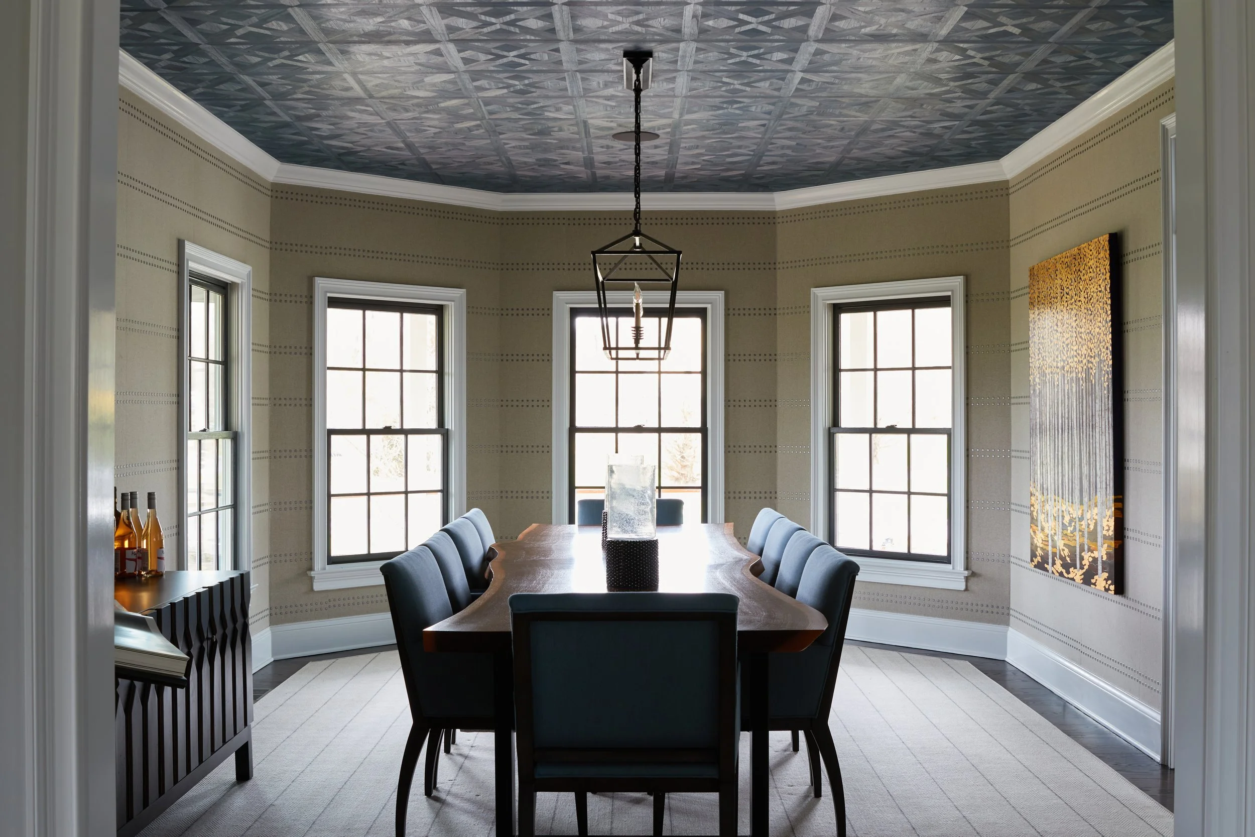 Dining room with a wooden table, eight chairs, three large windows, a modern light fixture, artwork on the wall, and a sideboard with bottles.