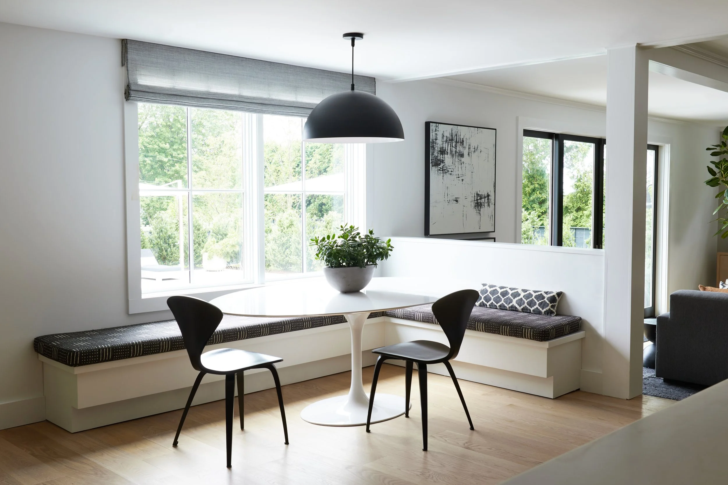 Eat-in breakfast nook with a white round table, two black chairs, a built-in window seat with black patterned cushions, a potted plant, large windows letting in natural light, and a modern black pendant light.