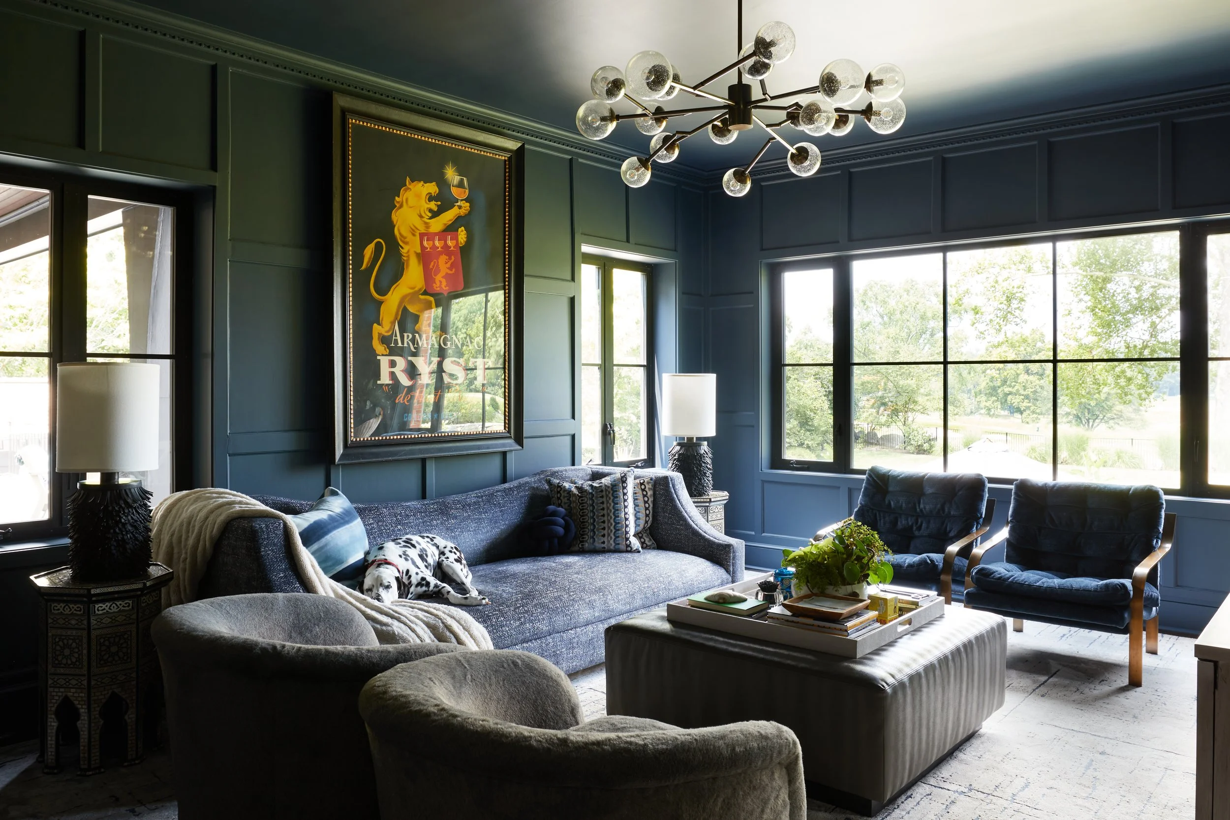 Living room with dark blue walls, large windows, a modern chandelier, a grey sofa with pillows, a Dalmatian puppy resting on a blanket, two armchairs, side tables with lamps, and a coffee table with plants and books.