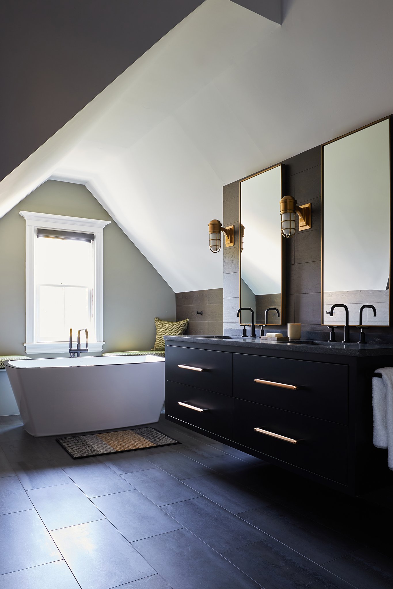 Modern bathroom with a black vanity, double mirrors, and two light fixtures, a white bathtub near a window, and gray tile flooring.