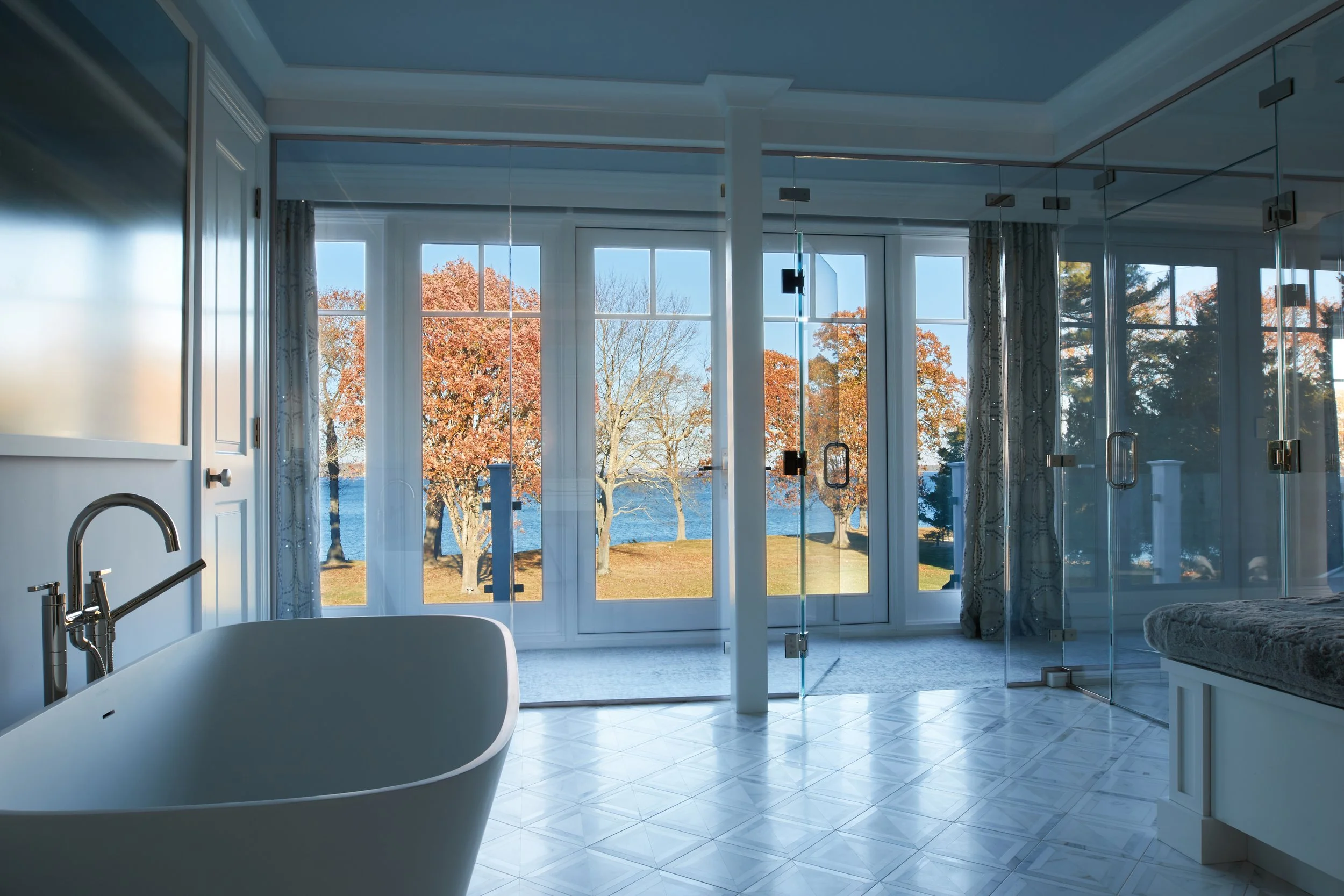 Modern bathroom with a freestanding bathtub near large windows featuring a view of trees and a lake, glass shower enclosure, tiled floor, and curtains.