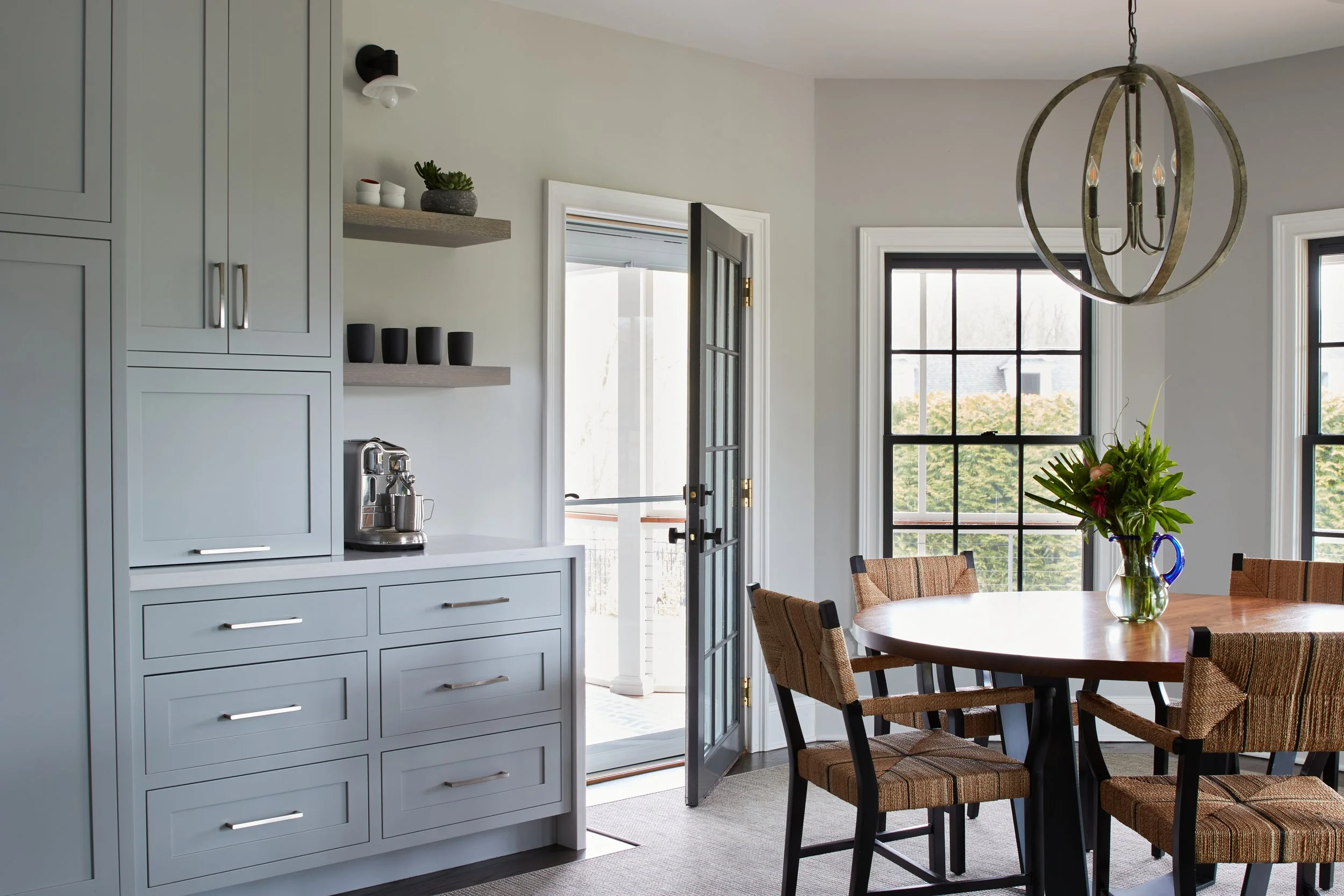 Dining room with large windows, round wooden table with flowers, and kitchen cabinets.
