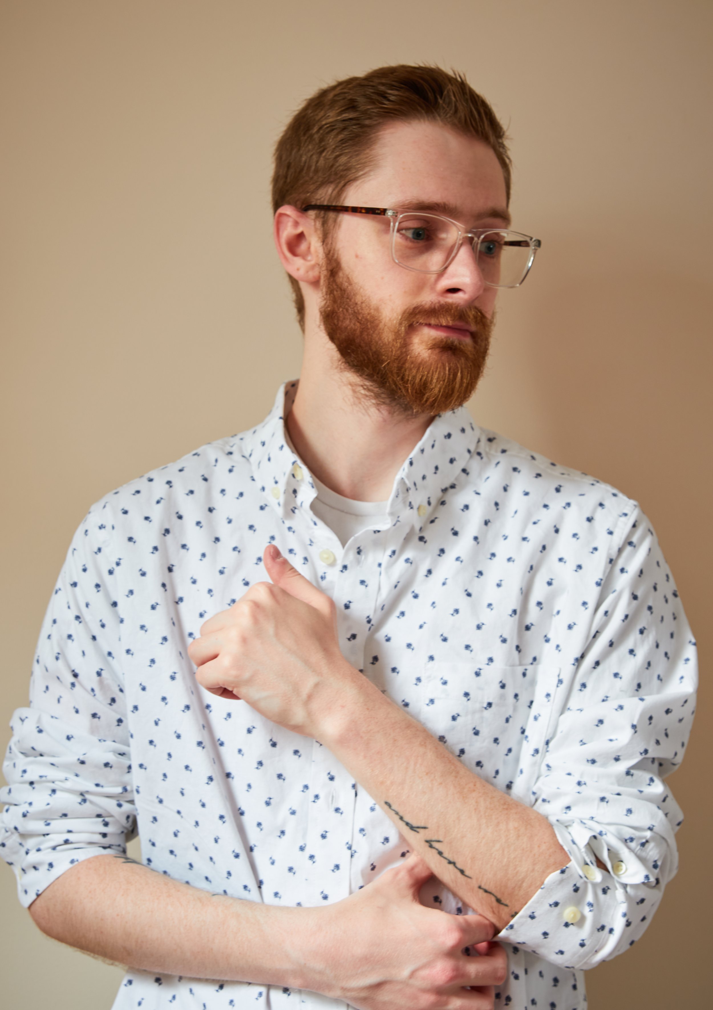 Joe Buzzeo wearing glasses and a white shirt with small blue patterns, looking down thoughtfully with one hand on his arm.
