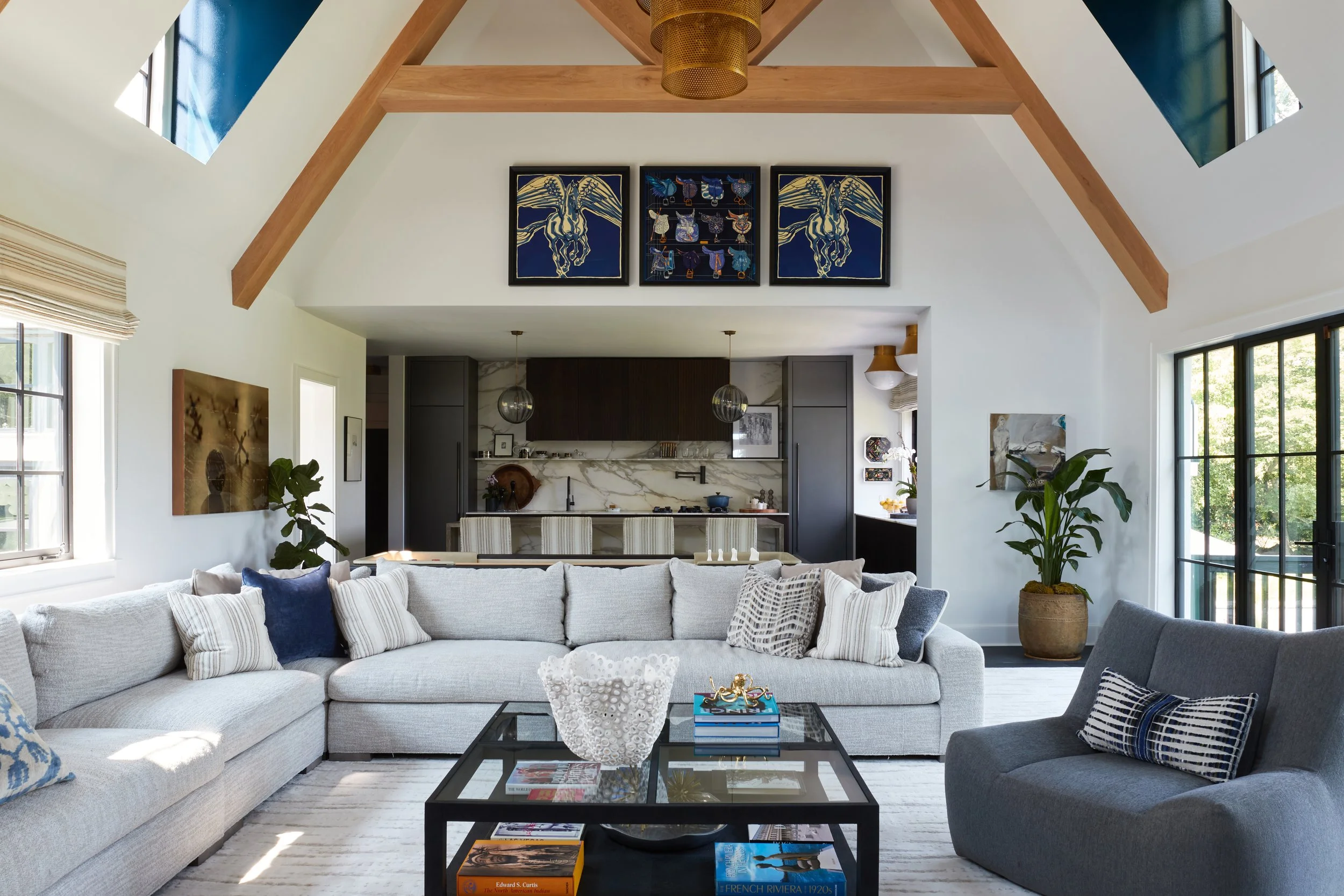 Living room with white sectional sofa, gray armchair, coffee table with books and decor, large windows, in an open floor plan with kitchen in the background, vaulted ceiling with wooden beams, modern art on walls, indoor plants, and natural light.
