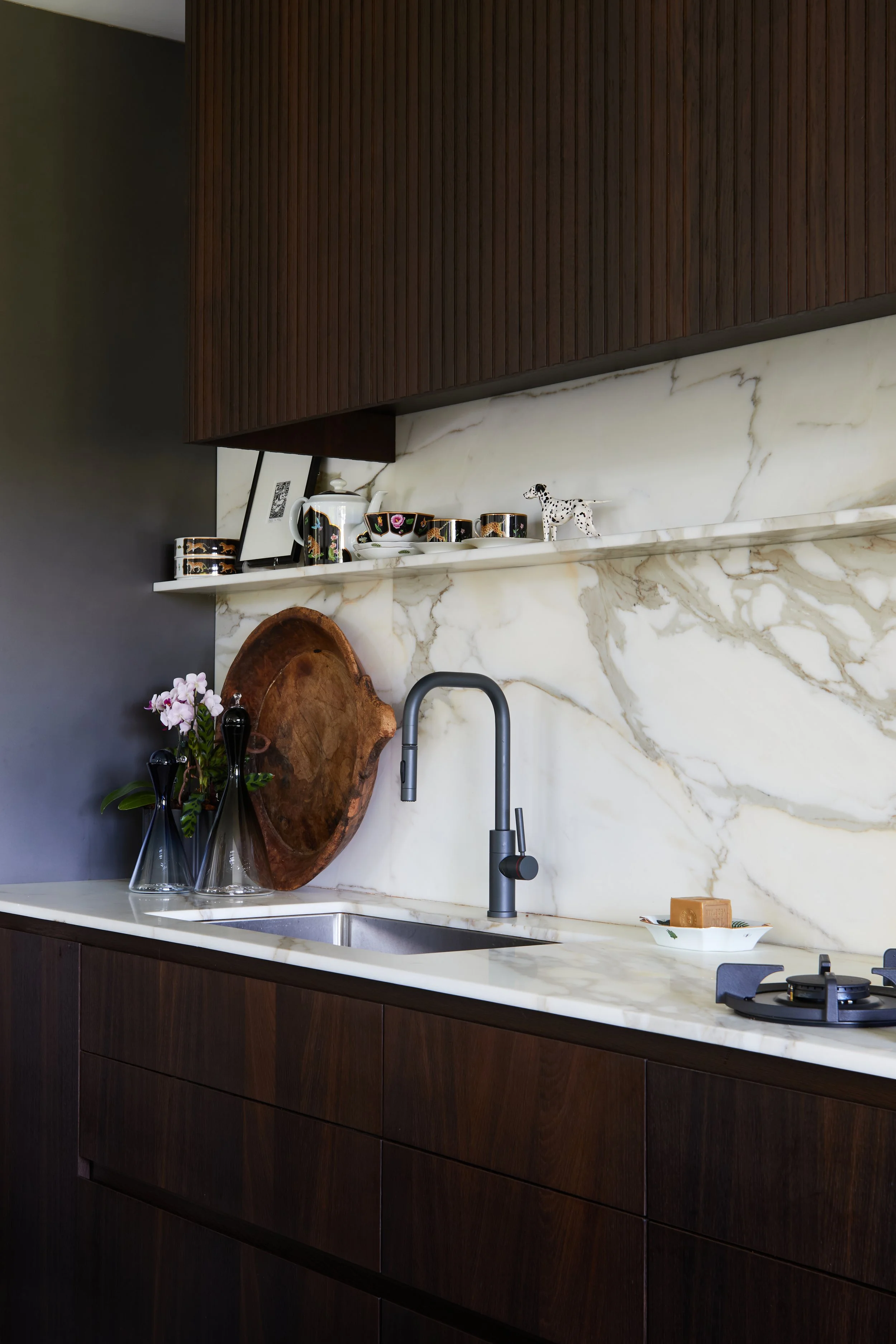 Modern kitchen with dark wood cabinets, white marble countertop and backsplash, black faucet, decorative bottles, flowers, and a soap bar.