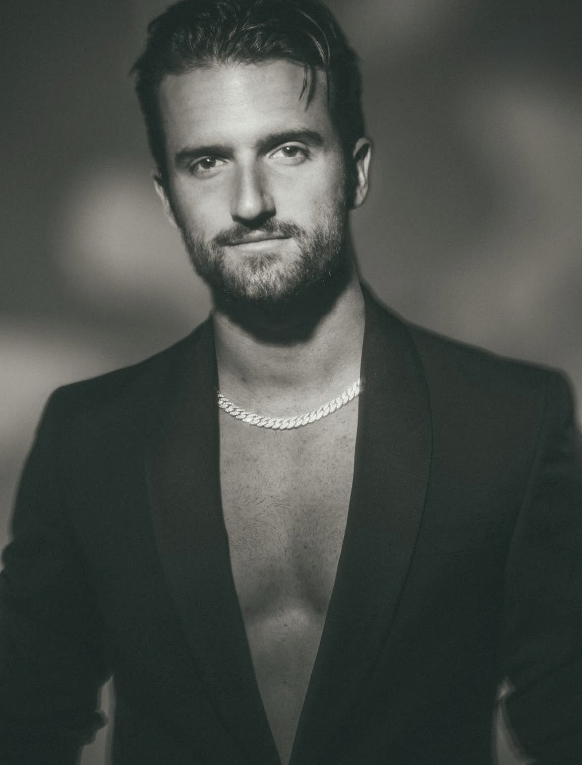 A black and white portrait of a man with a beard, short hair, partially unbuttoned blazer revealing chest hair, and wearing a chain necklace.