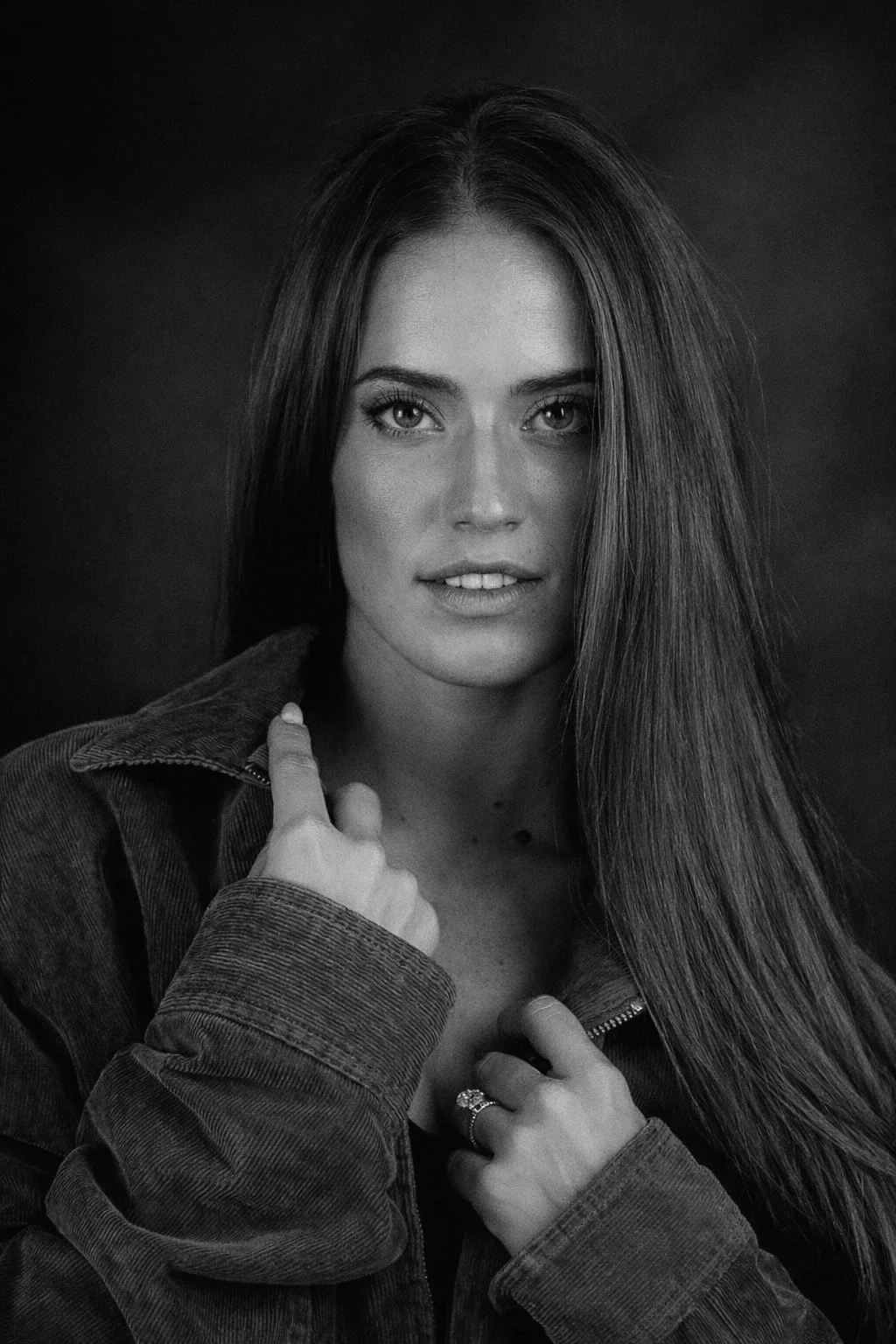 Black and white portrait of a woman with long straight hair, wearing a denim jacket, looking at the camera with a slight smile, against a dark background.
