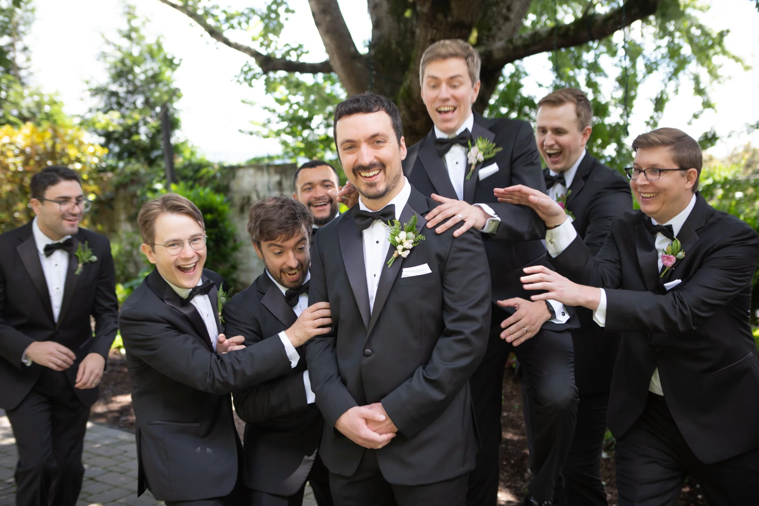 A group of groomsmen in tuxedos with boutonnières laughing and smiling in an outdoor garden setting with trees and greenery.