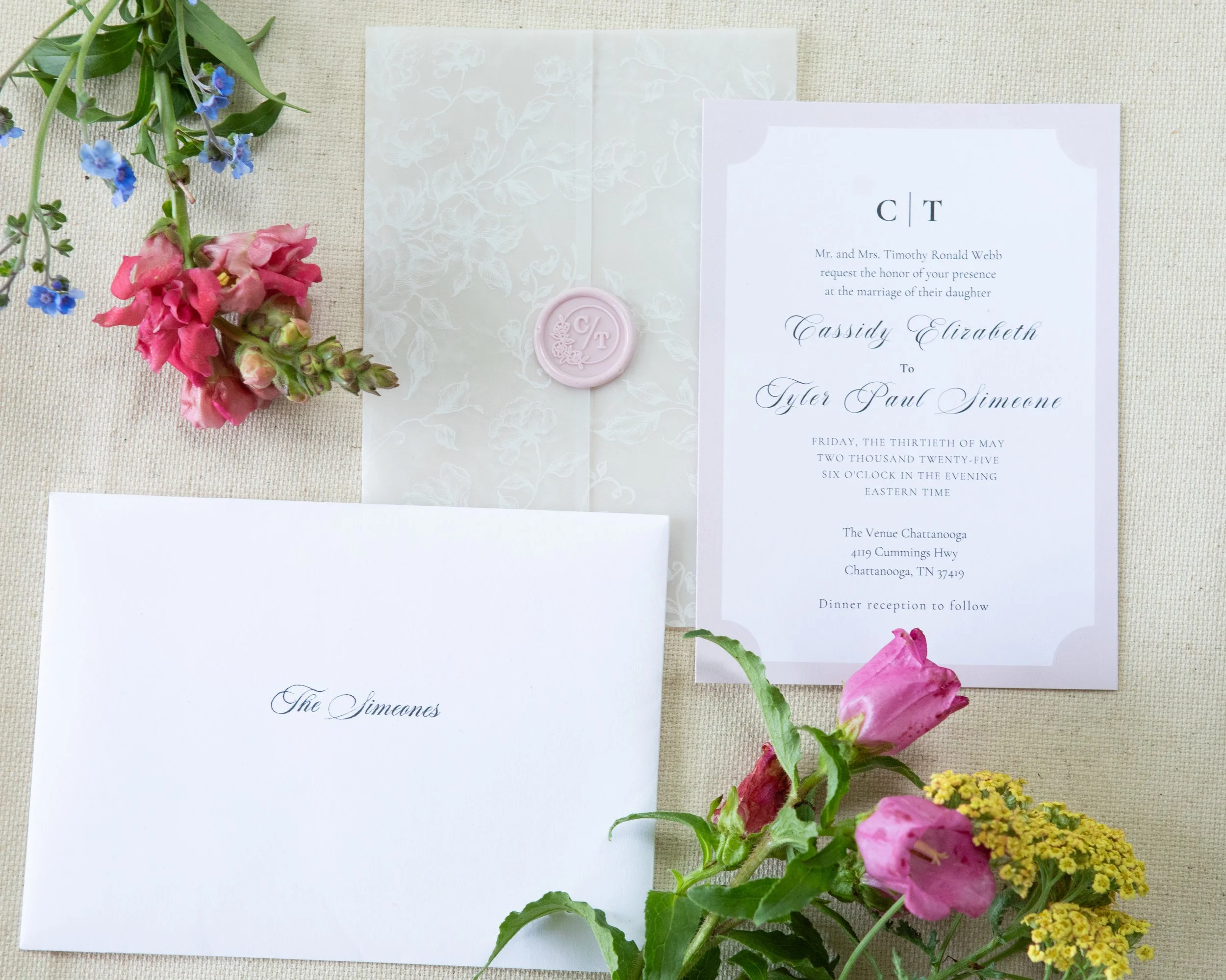Wedding invitation suite with a white envelope, a detailed white patterned translucent paper, and a ceremony invitation card, decorated with pink roses, purple flowers, yellow flowers, and a pink wax seal on the paper.